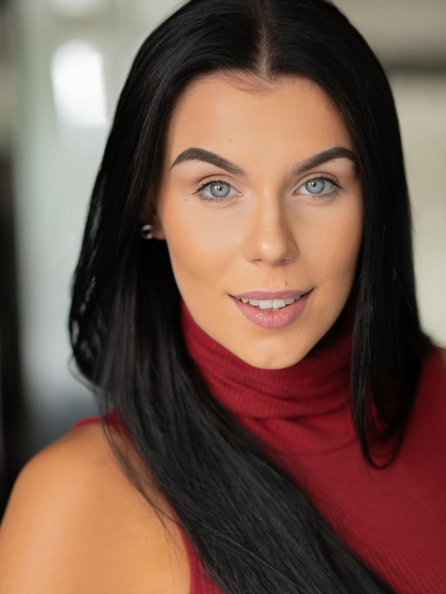 Close-up portrait of a young woman with long black hair, blue eyes, wearing a red turtleneck, smiling softly indoors.