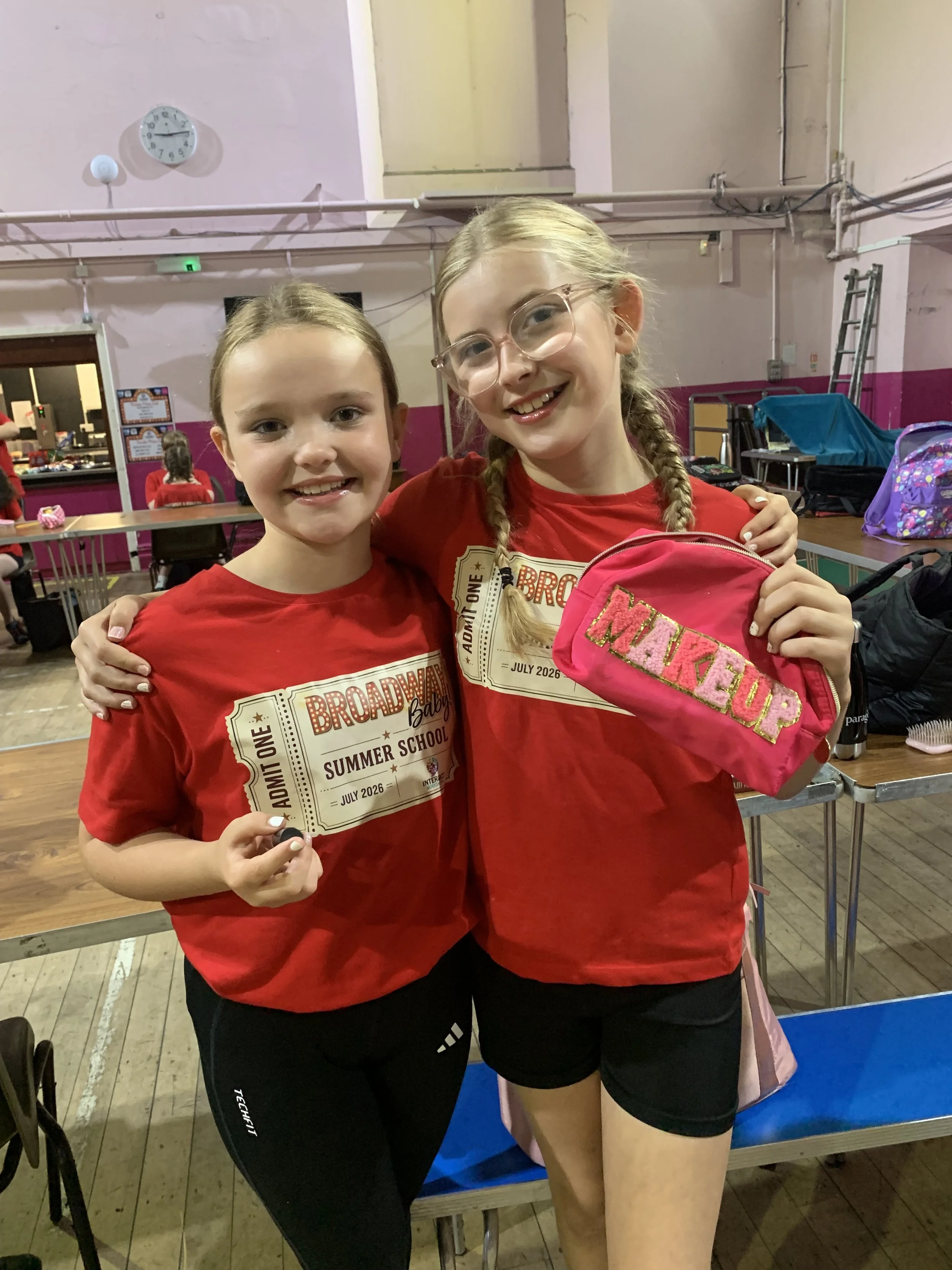 Two young girls smiling, wearing red shirts, holding a pink bag labeled 'MAKE UP' in a room with pink walls, tables, and chairs.