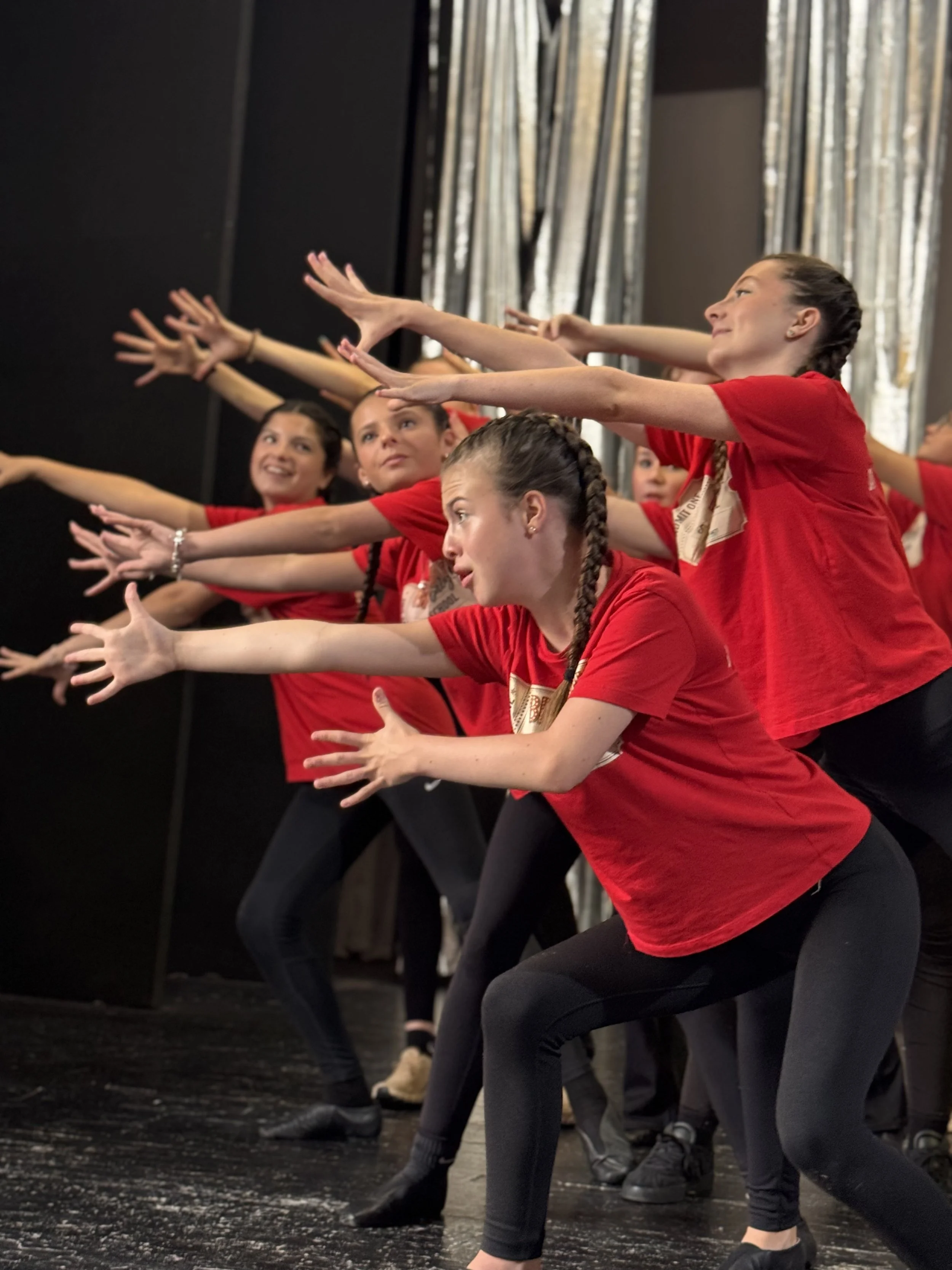 Group of young women in red shirts and black leggings practicing dance moves on stage, stretching with arms extended forward and focused expressions.