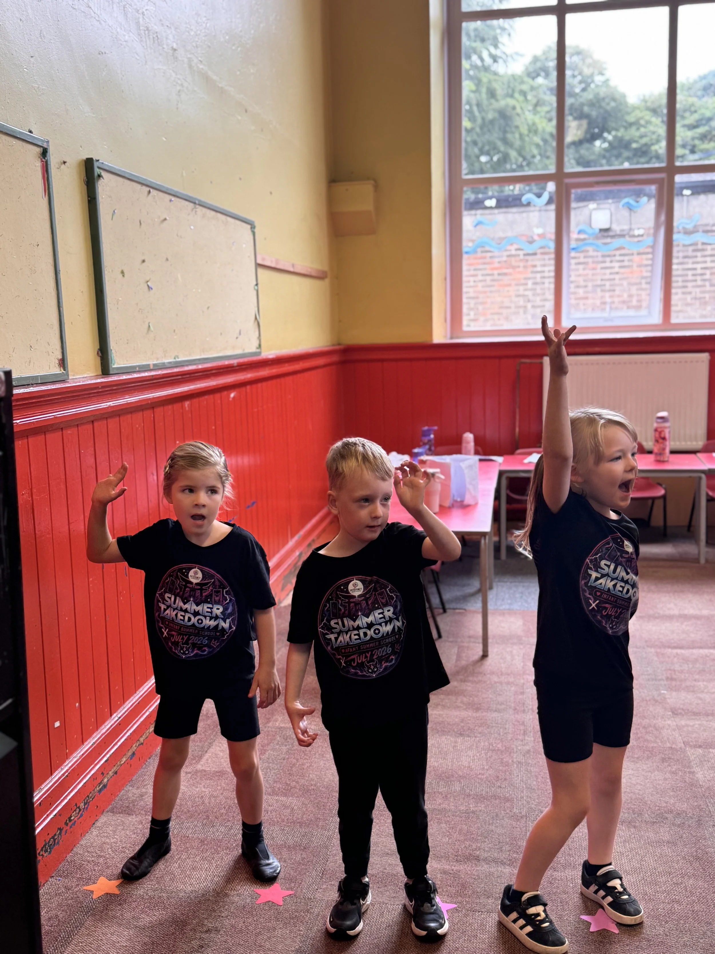 Three children wearing black 'Summer Takedown' t-shirts standing in a room with red wainscoting and a large window, raising their hands, with colorful star-shaped paper decorations on the floor.