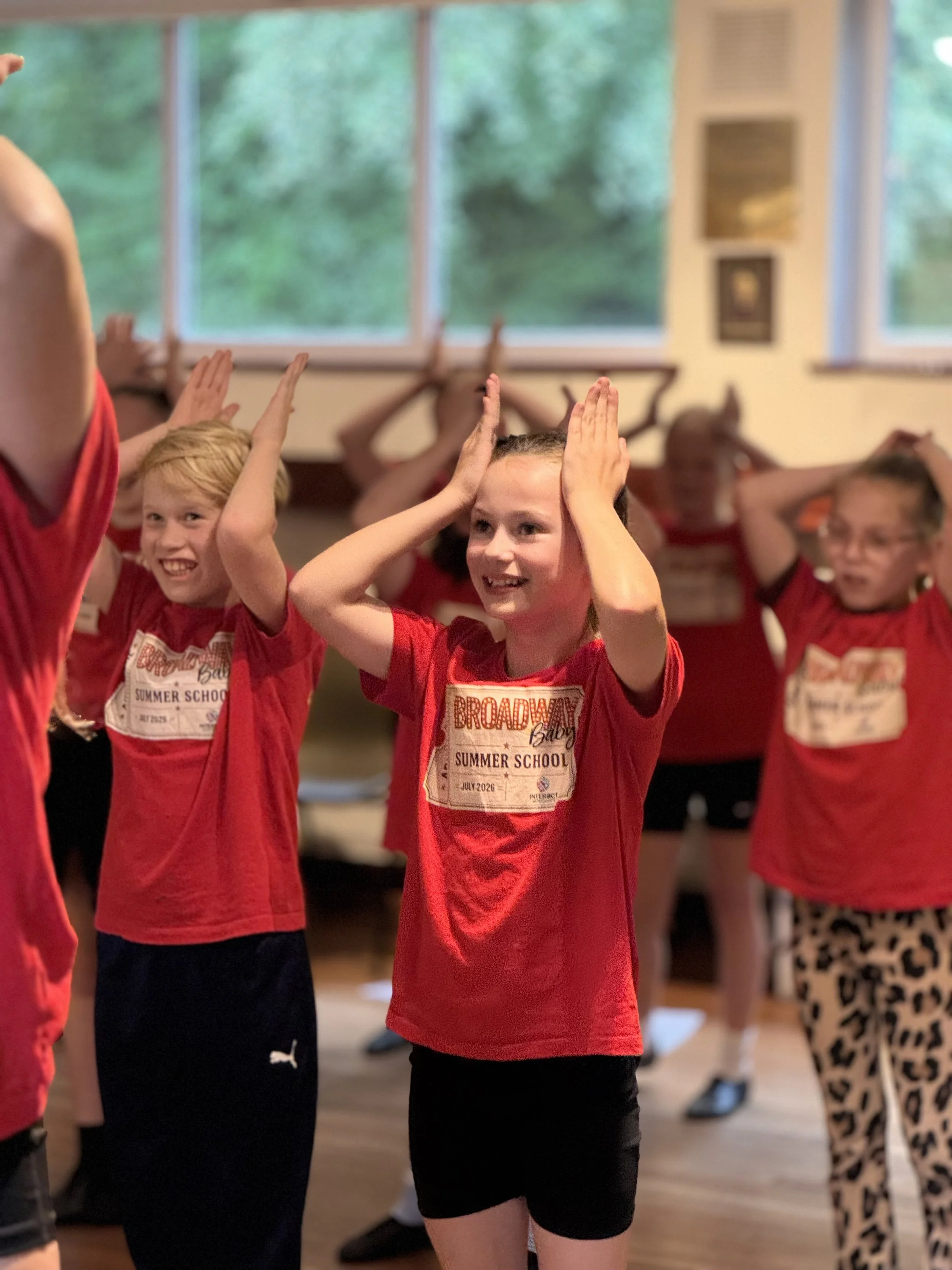 Children participating in a dance or movement activity inside a room, wearing red T-shirts with 'Broadway Baby Summer School' printed on them.