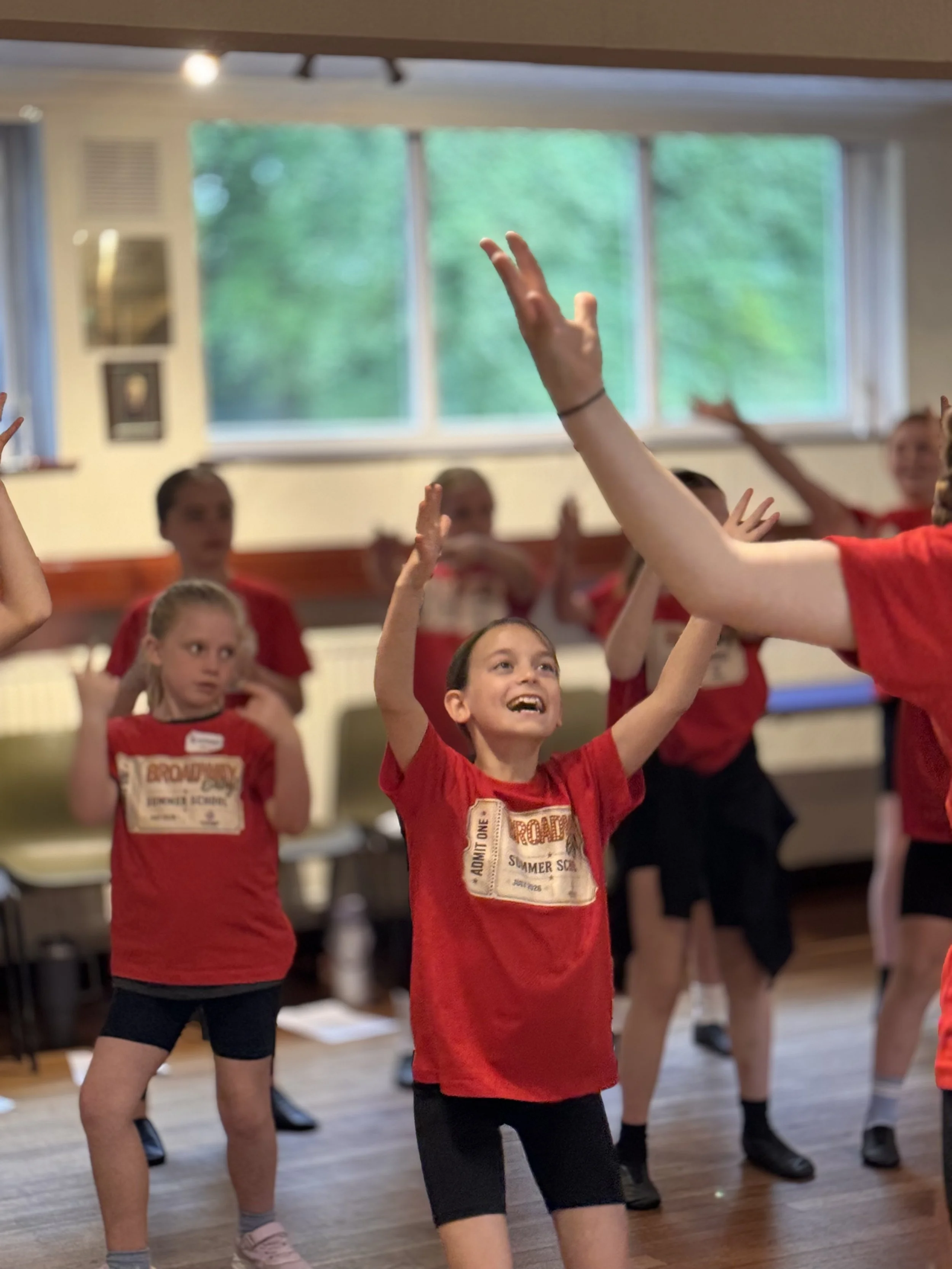 Children participating in a dance or exercise class inside a room with large windows, wearing red t-shirts and black shorts, raising their hands.