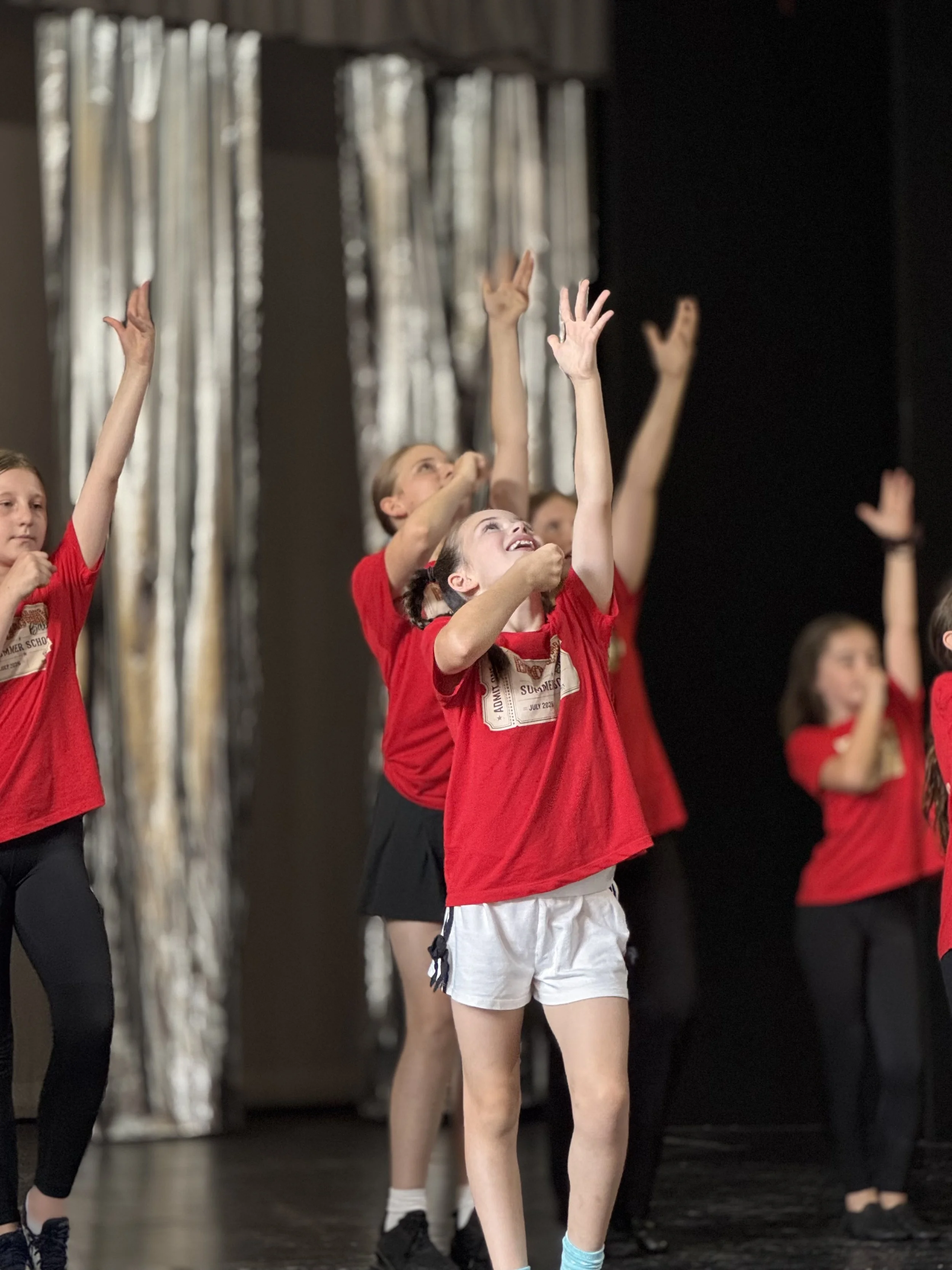 Children in red shirts and shorts performing a dance on stage, raising their arms with joyful expressions.