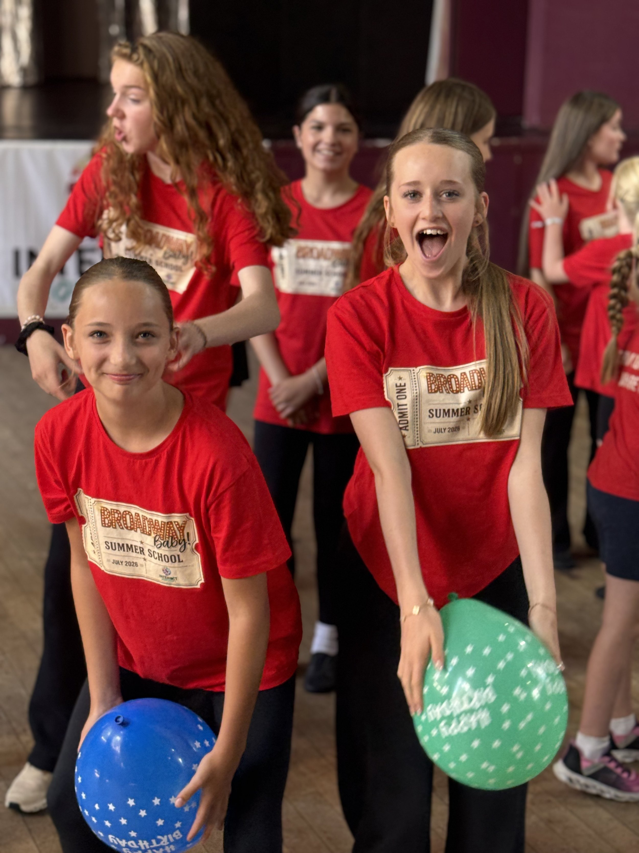 Group of young girls in red shirts holding colorful balloons at an indoor event.