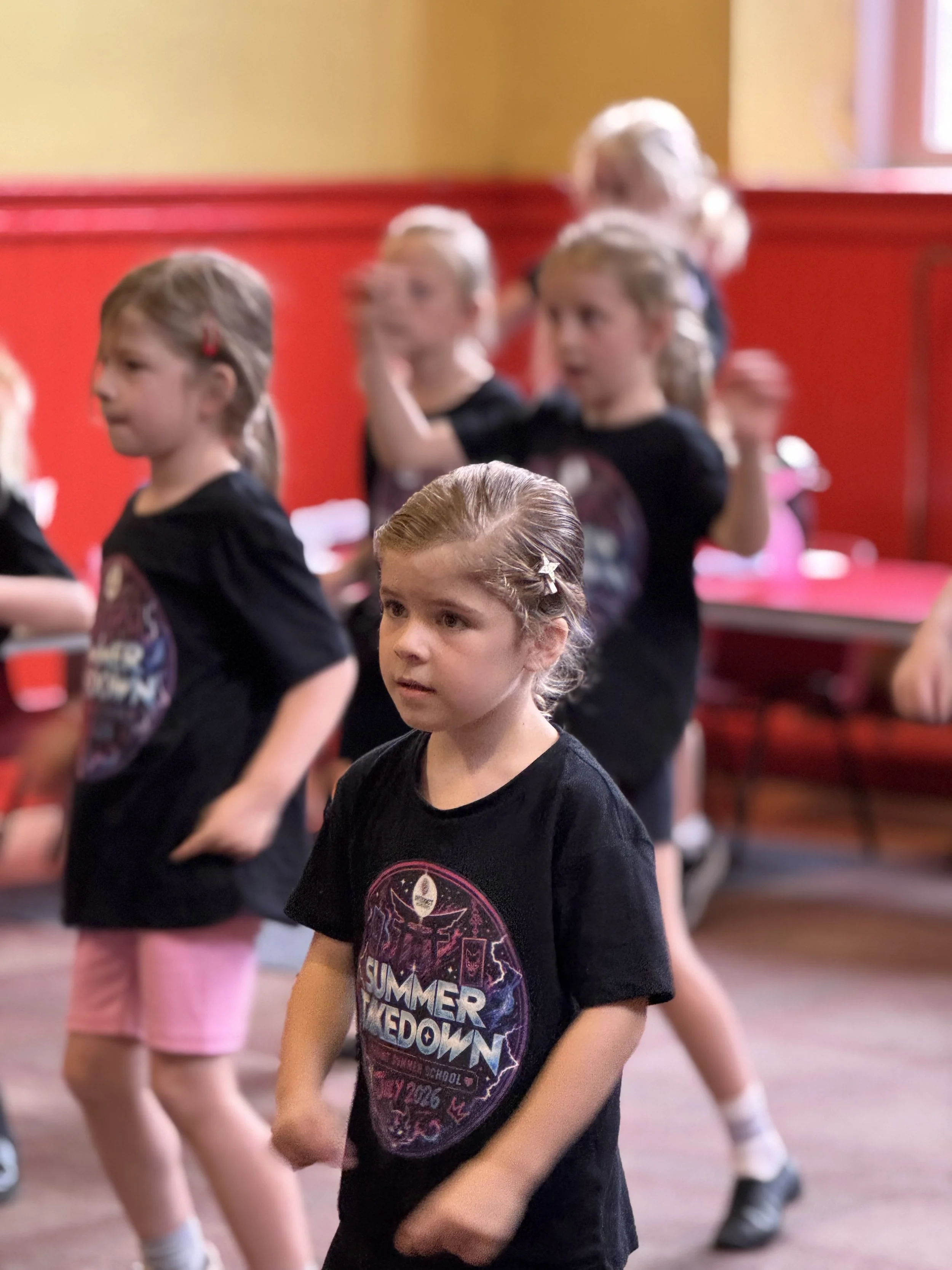 Young girls participating in a dance class or rehearsal, wearing black t-shirts and pink shorts, in a room with yellow and red walls.
