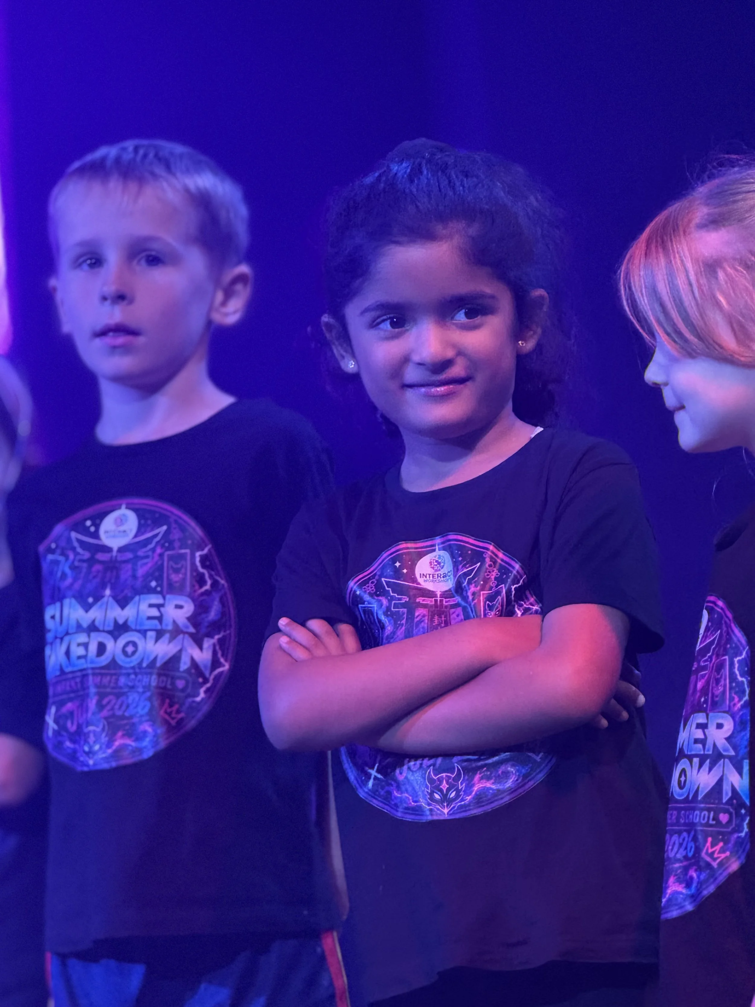 Three kids in matching black shirts with colorful designs, standing in a dark, possibly neon-lit environment. The girl in the middle has black hair styled in a ponytail, with her arms crossed and smiling slightly. The boy on the left has short light 