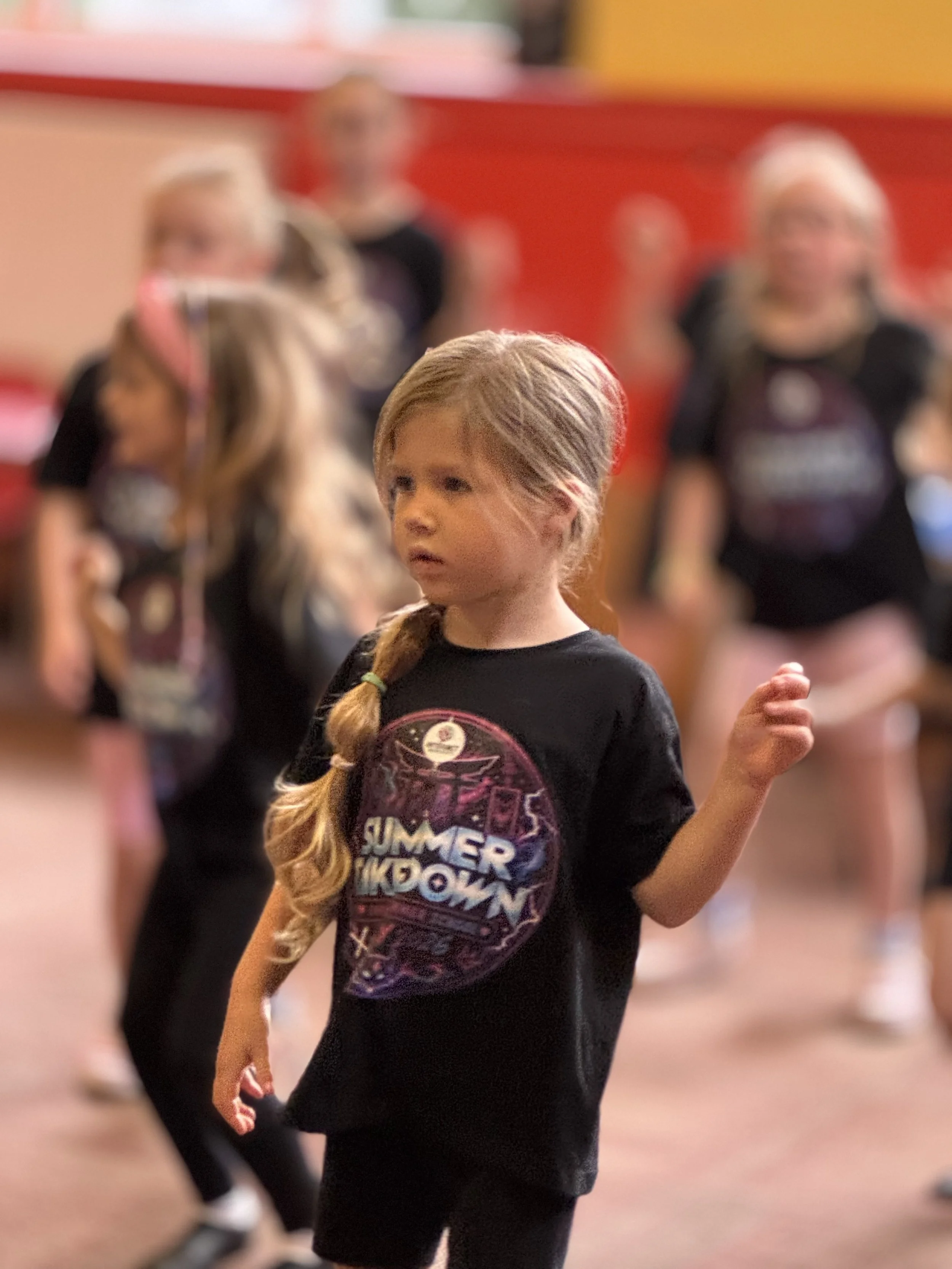 A young girl with a braid wearing a black t-shirt with a colorful graphic design, standing in a room with other people blurred in the background.