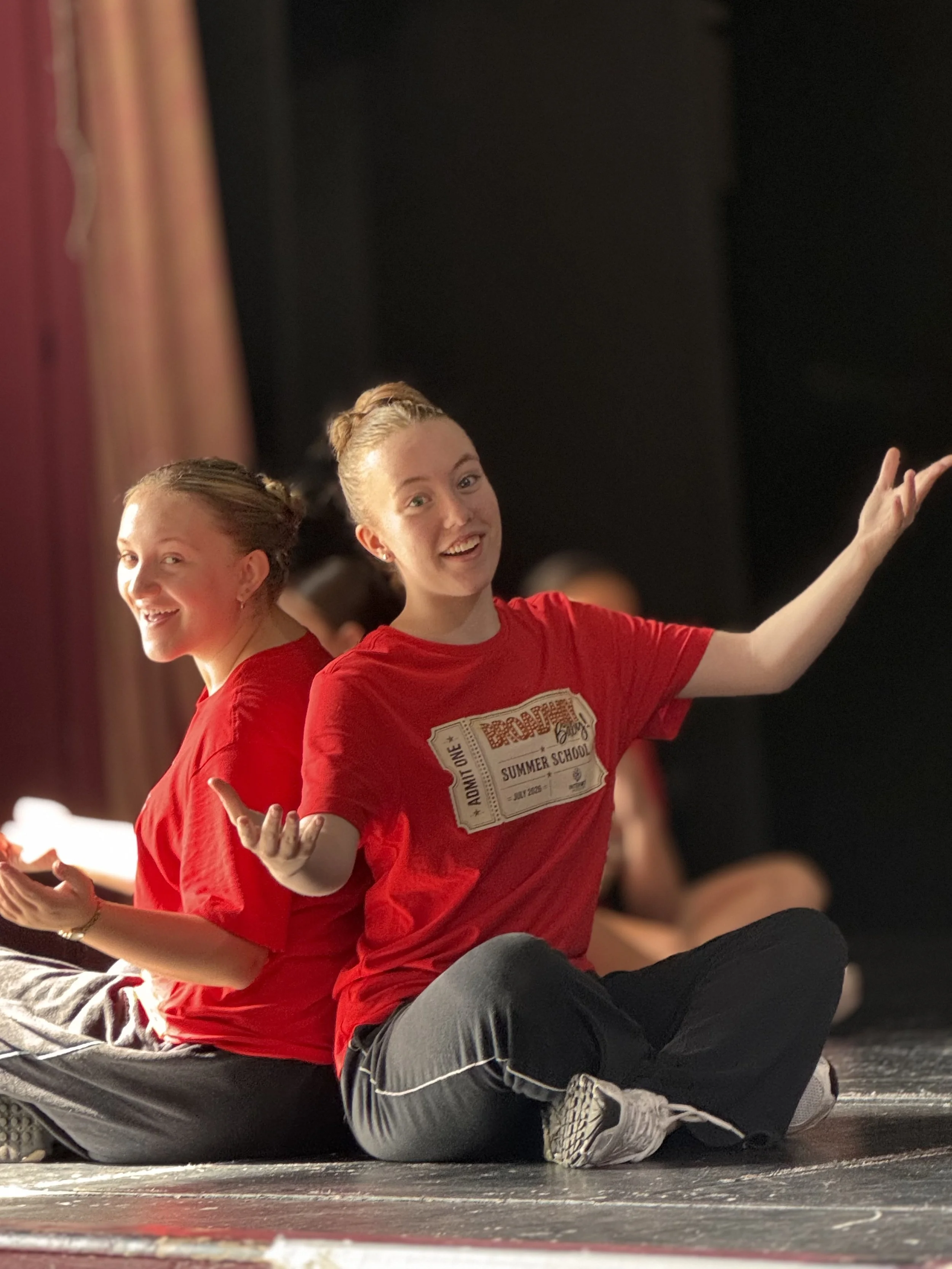 Two young women dressed in red shirts sitting on the floor, smiling and posing for the camera with arms extended. They are in a dimly lit room with a dark background and a curtain to the left.