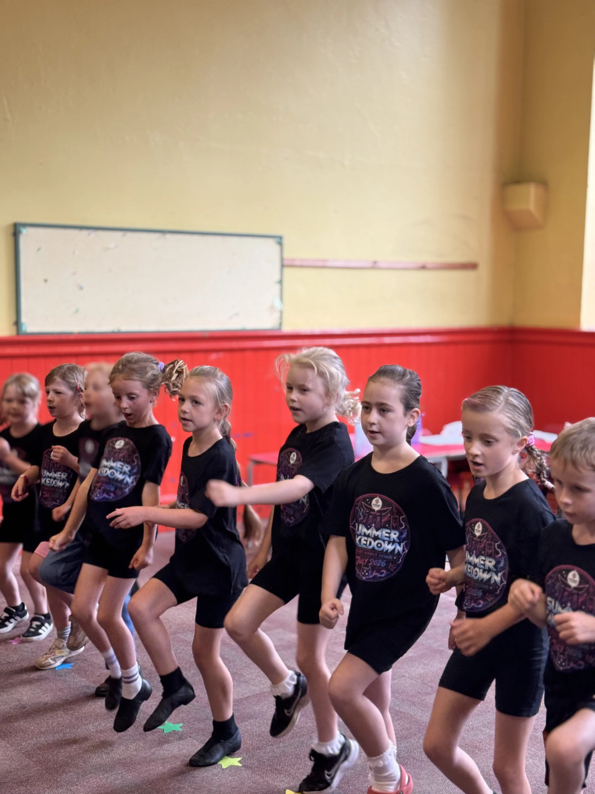 Group of young girls with ponytails participating in a dance or exercise class in a gymnasium, wearing black T-shirts with a colorful logo, black shorts, and sneakers, walking in a line with arms outstretched.