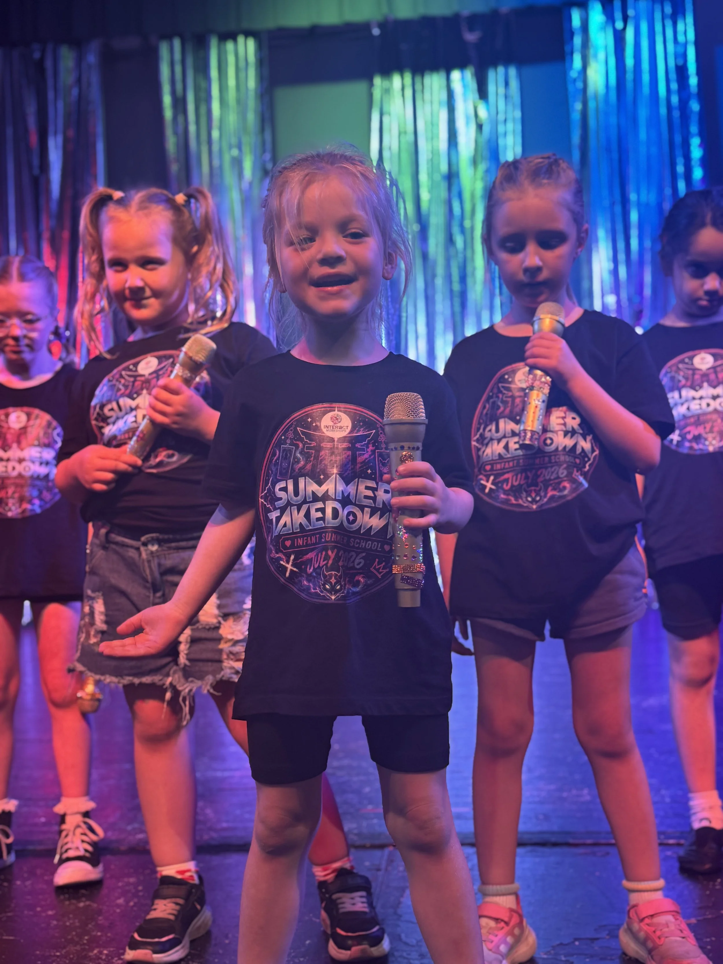 Young girls performing in a stage production wearing black T-shirts with colorful graphics, holding microphones, standing in front of a shiny, multicolored background.