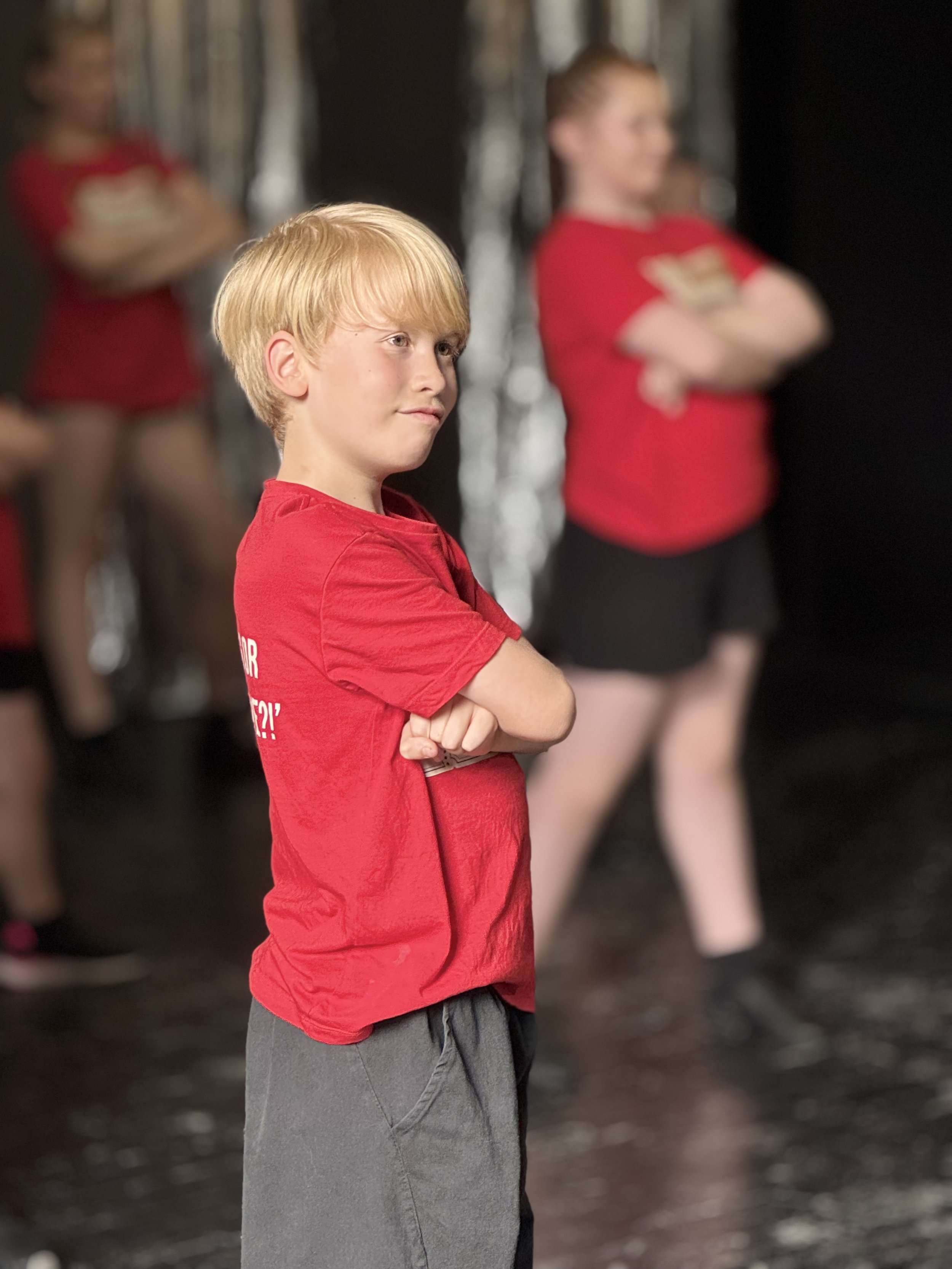 A young boy with blonde hair wearing a red shirt and gray shorts standing with crossed arms, with two other children in red shirts blurred in the background.