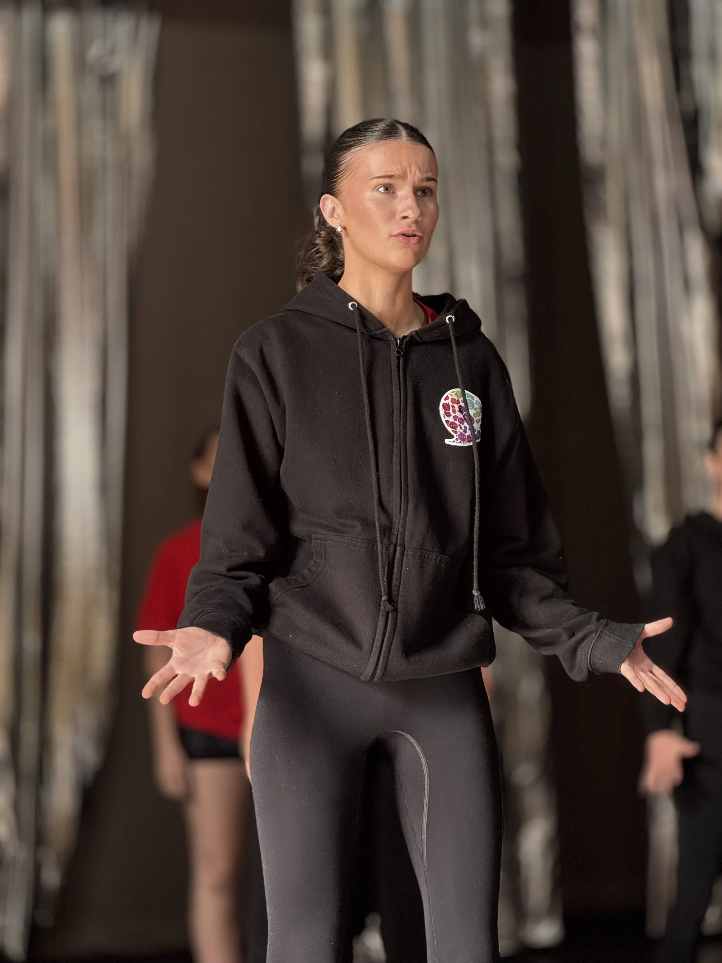 A woman wearing a black hoodie and black leggings, with her hair pulled back, standing with her hands open and making a gesture, in a room with clothing racks and other people in the background.