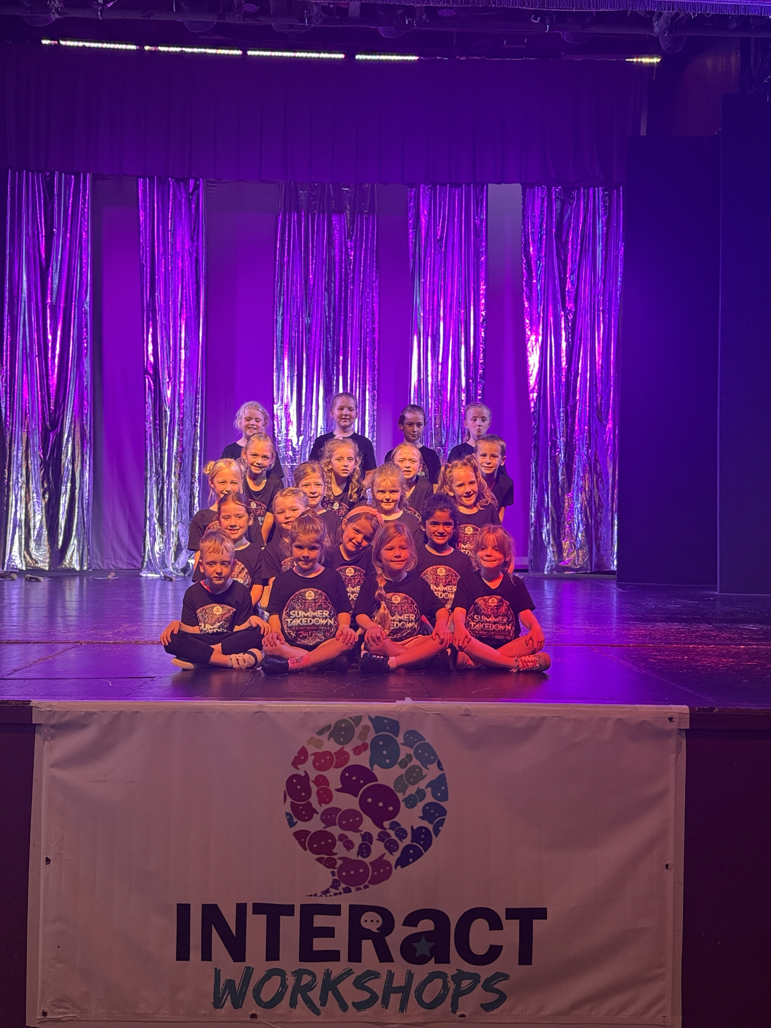 A group of children on stage for a performance at the INTERACT Workshops event, with a purple curtain backdrop and a banner in front displaying the event name and logo.