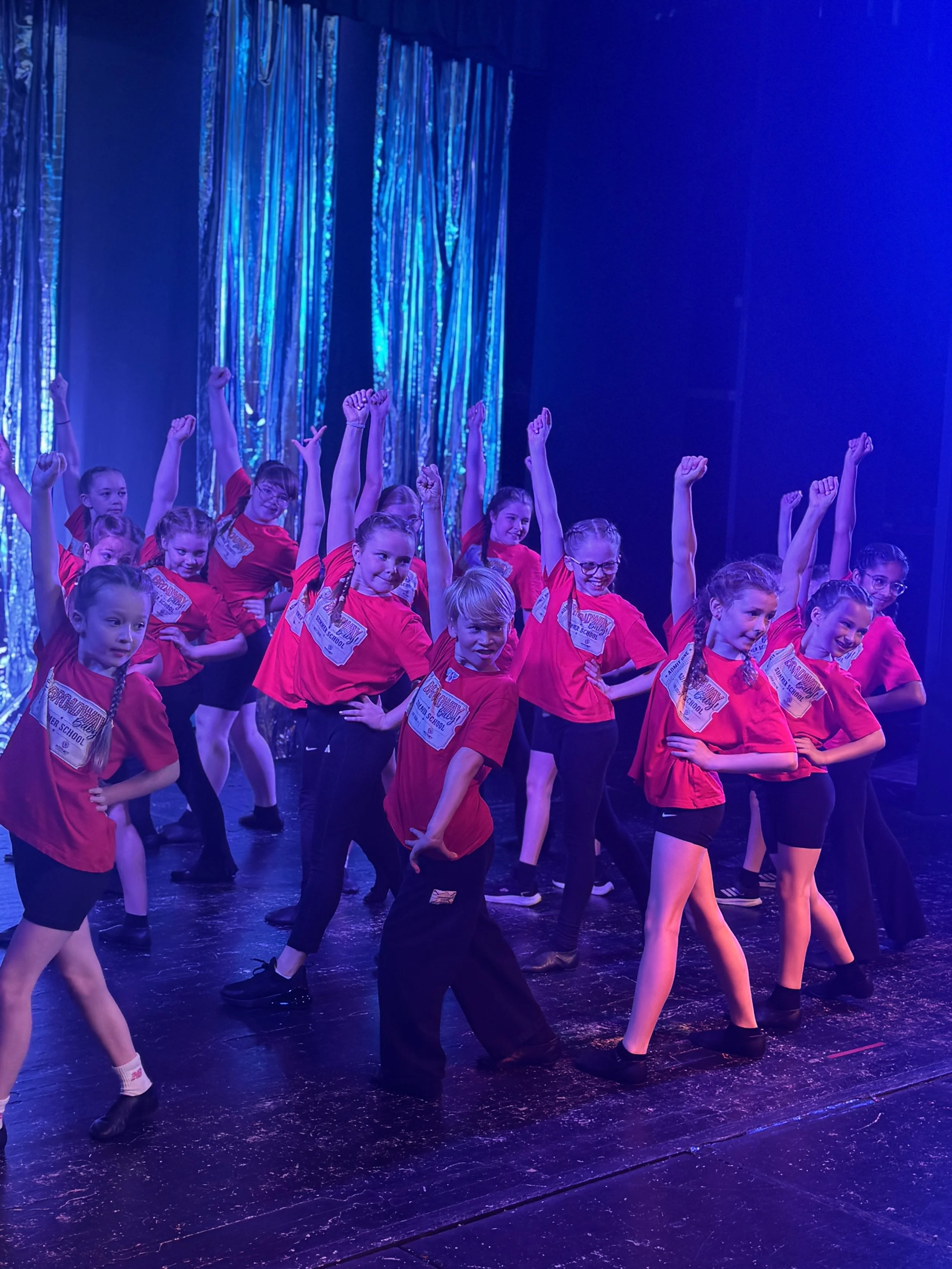Young girls performing a dance routine on stage, wearing red shirts and black bottoms, with some raising their arms and smiling. The background features shiny blue streamers and a dark stage floor.