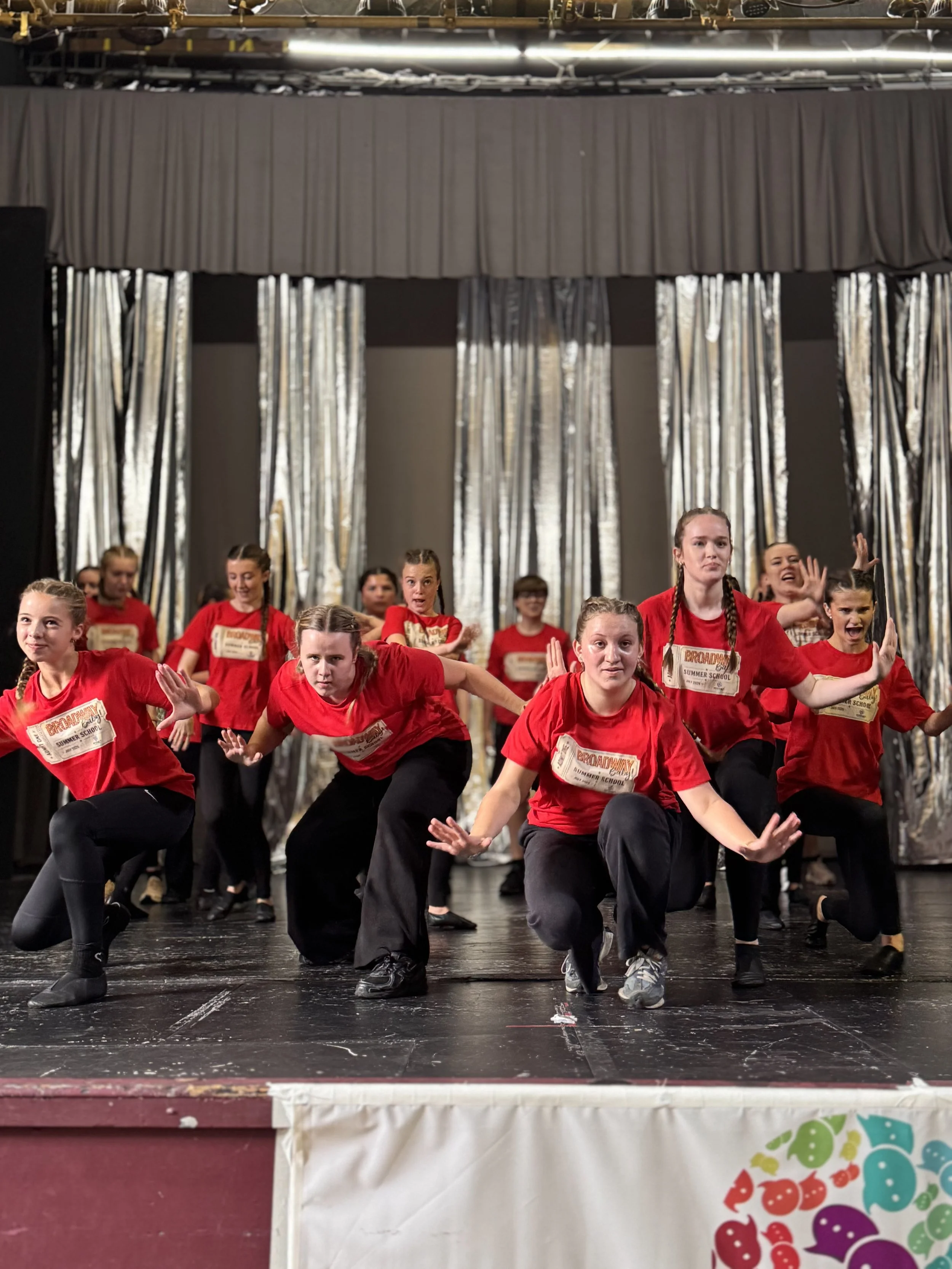Group of children performing a dance routine on stage, dressed in red shirts and black pants. They are in various dance poses with focused expressions. The stage has a black floor and curtains in the background. There is a colorful banner at the fron