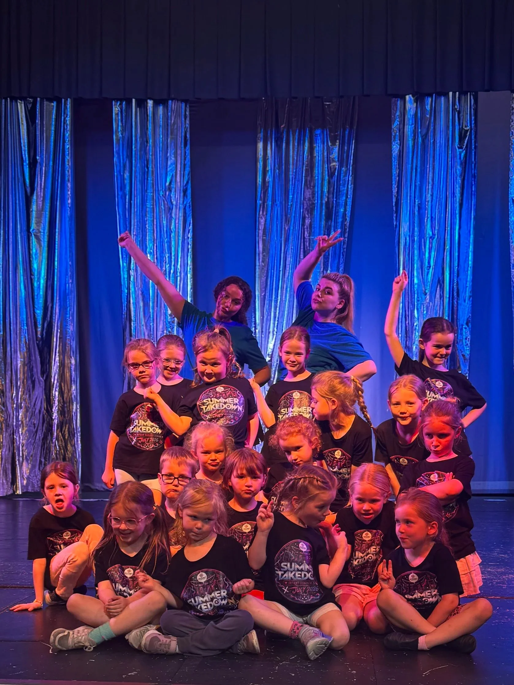 Group of children and young adults on stage during a dance performance, wearing black t-shirts with 'Summer Takedown' written on them, against a blue curtain backdrop with shiny metallic curtains.