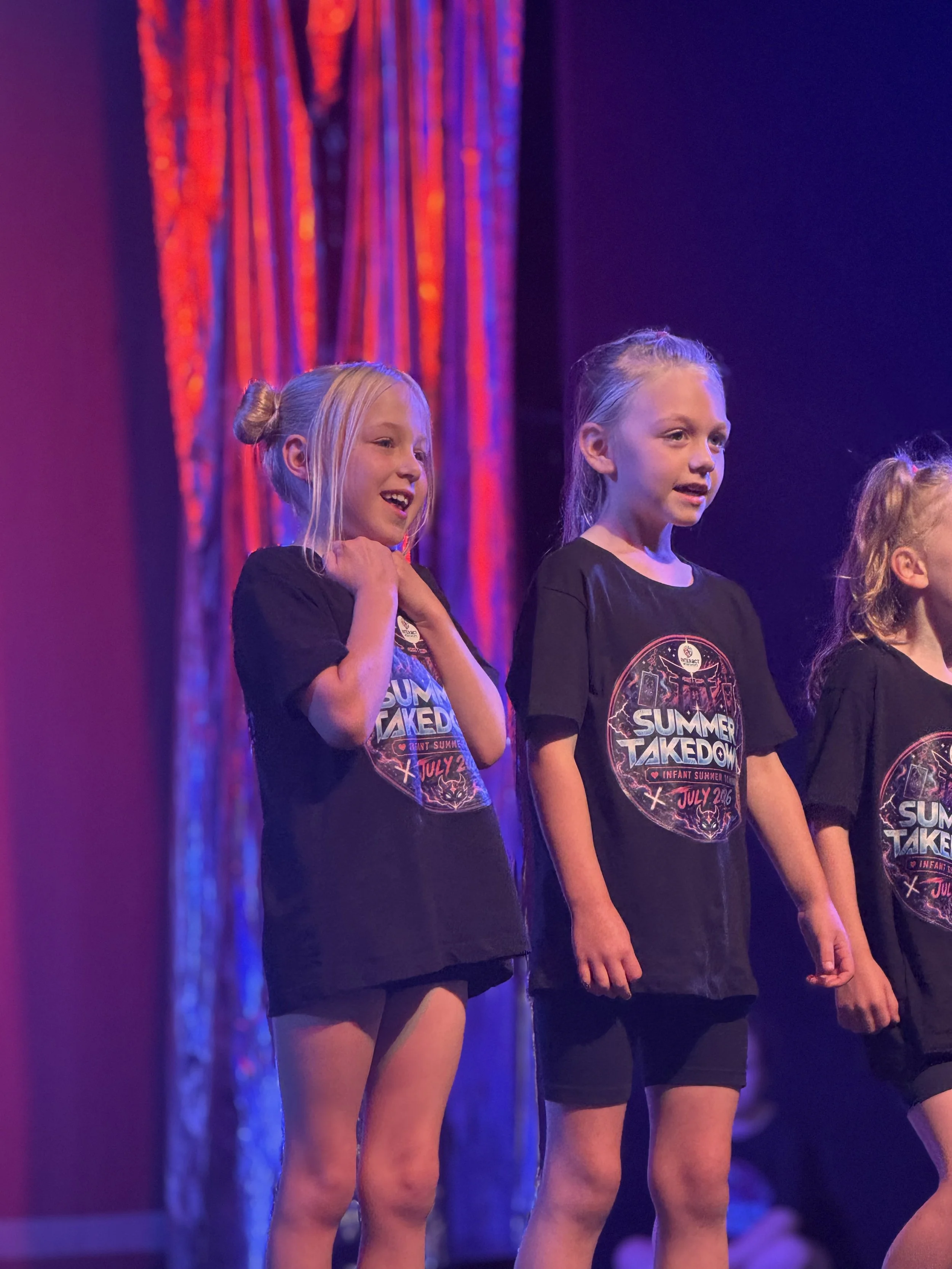 Three girls standing on stage with a red curtain in the background, wearing black T-shirts with a summer event logo, possibly during a talent show or performance.