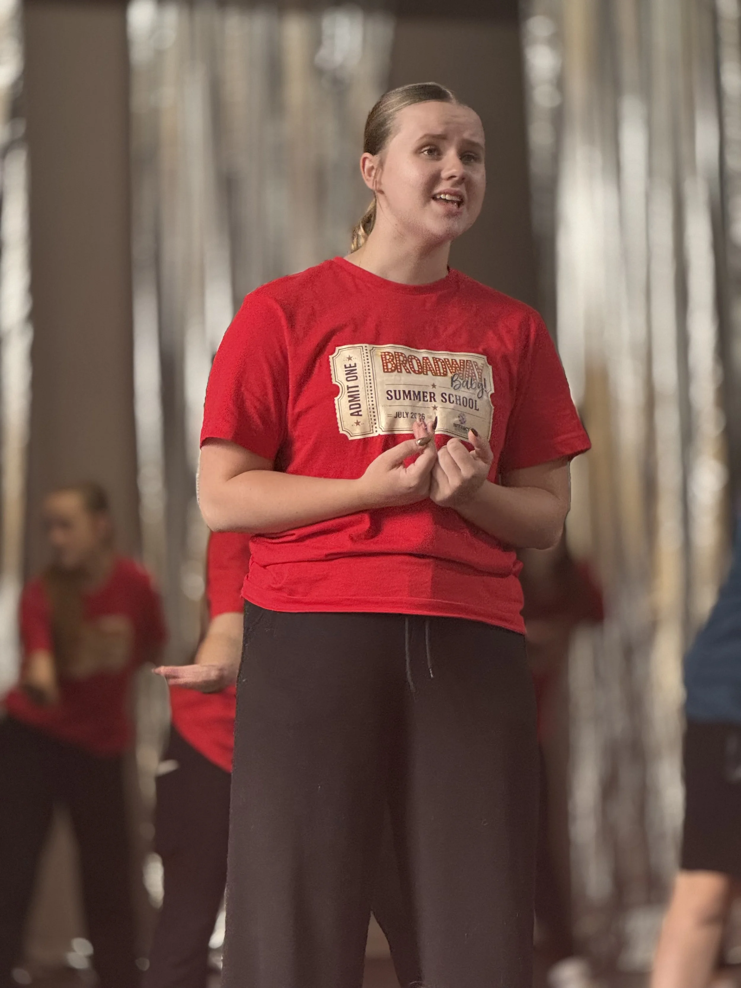 A woman in a red shirt with a Broadway Summer School logo on it is standing among a group of people.