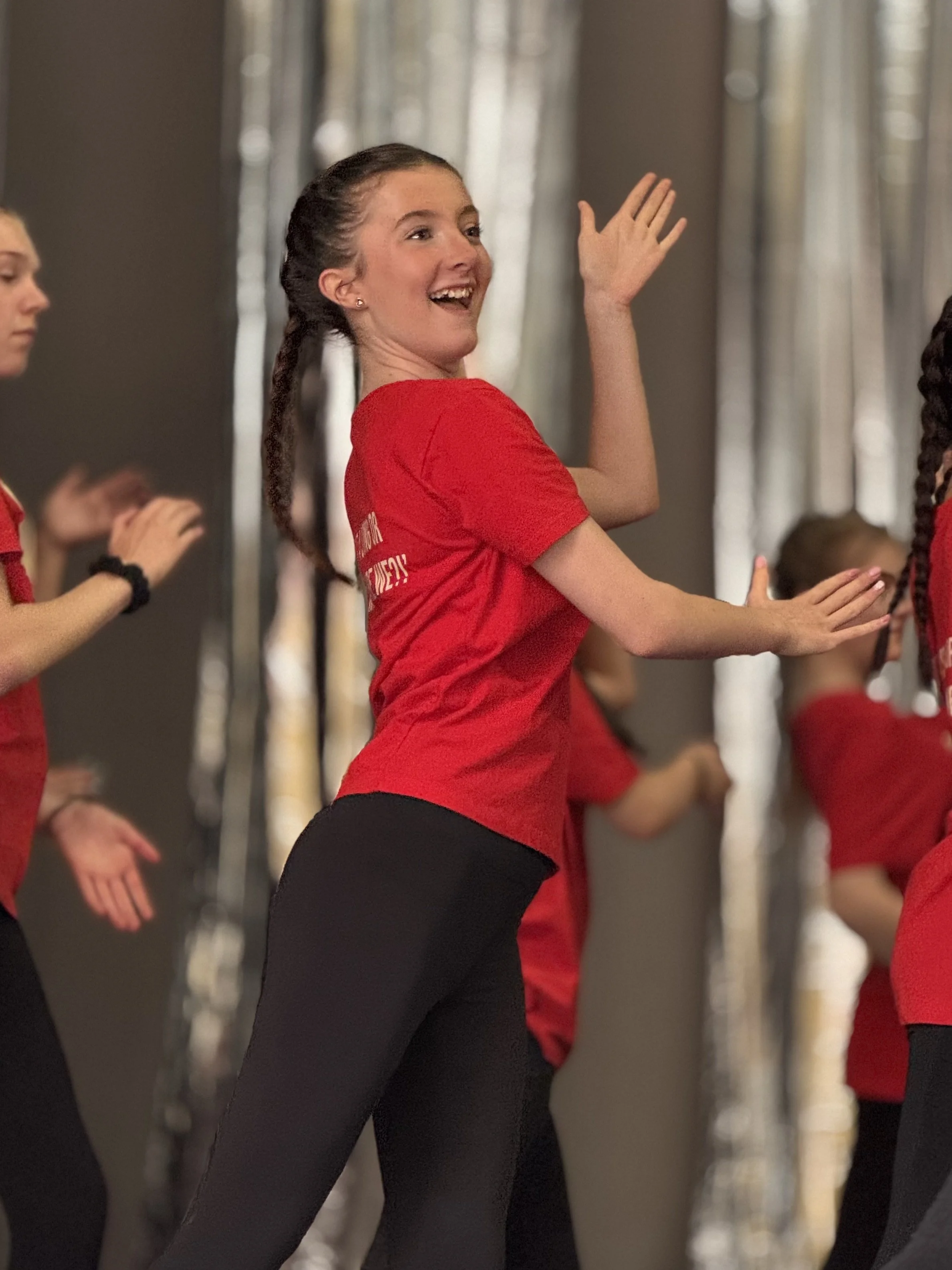 A group of young women participating in a fitness class, wearing red shirts and black leggings, standing in a line with synchronized arm movements.