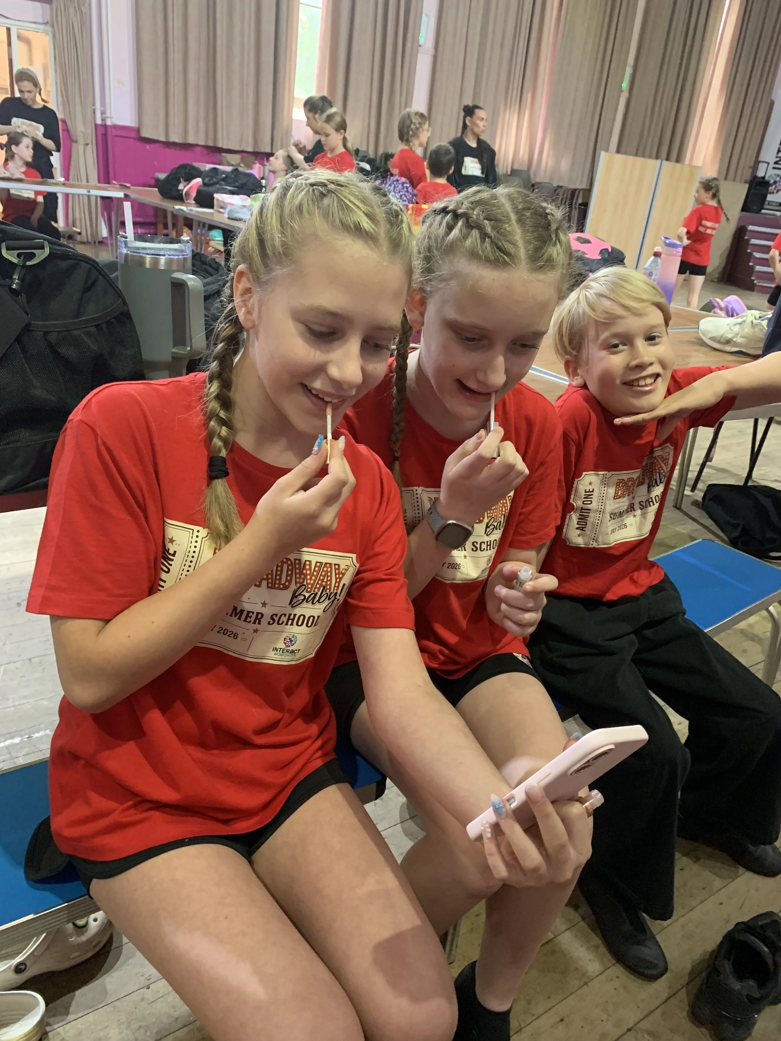 Three young girls in matching red shirts sitting on chairs, looking at their phones, with one girl smiling at the camera, inside a room with other children and adults in the background.