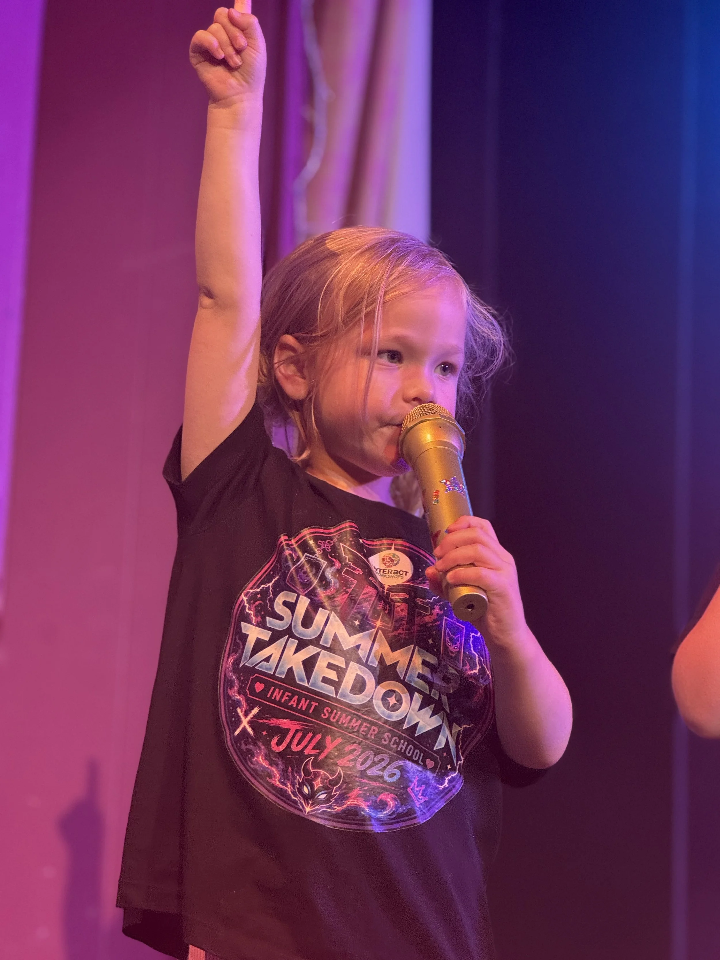 A young girl with blonde hair holding a gold microphone with a star decoration, wearing a black t-shirt that reads 'Summer Takedown,' at a summer school event in July 2026 with purple and pink lighting.