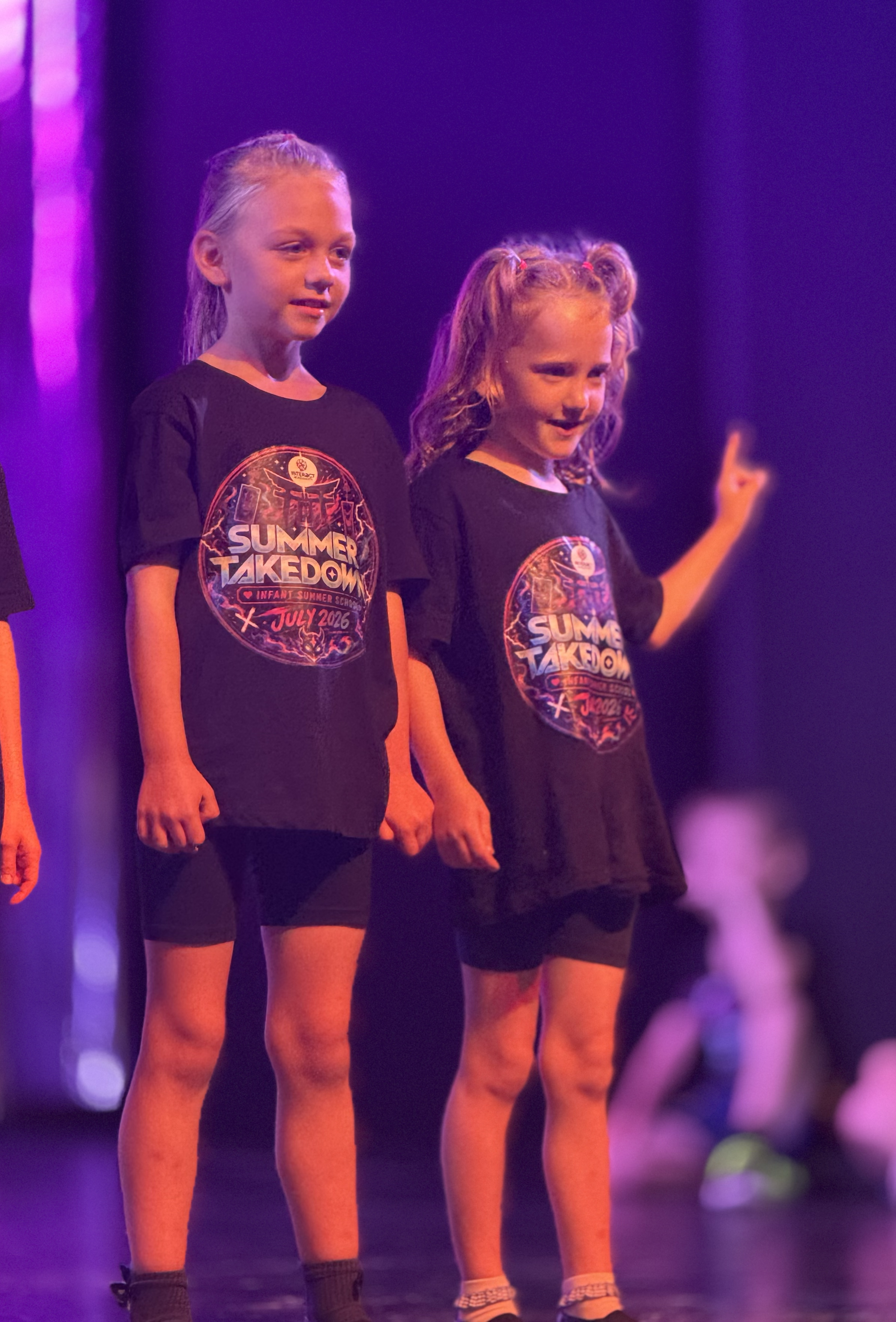 Two young girls wearing black T-shirts with a colorful 'Summer Takedown' logo, standing on stage with purple lighting behind them, lips slightly parted, looking down, with their hands at their sides.