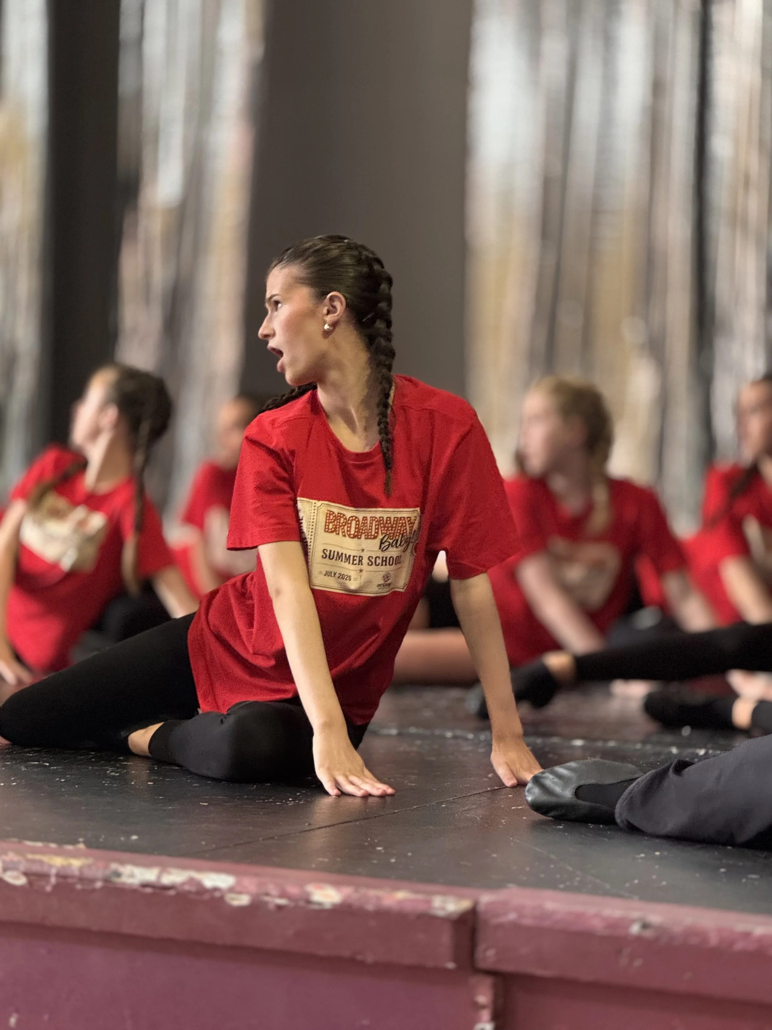 A woman in a red shirt performing a stretch or yoga pose on a black mat, with others in red shirts sitting or stretching behind her in a studio or gym.