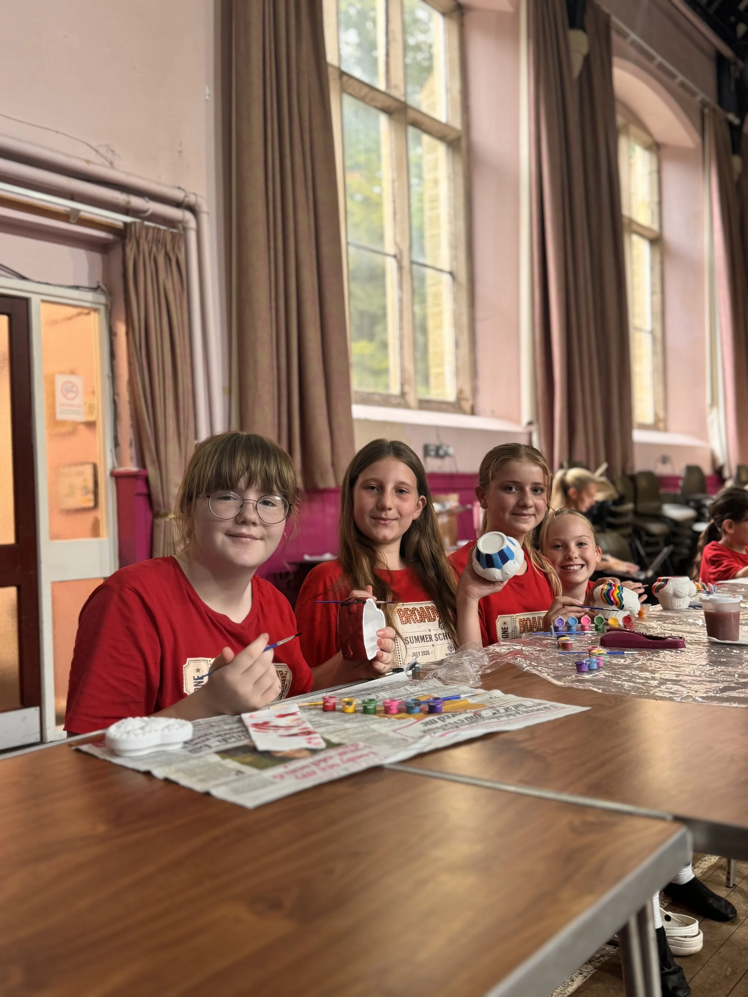 Group of young children sitting at a table in an indoor setting, participating in a craft activity with painted pottery and art supplies. They are wearing red shirts and smiling. The room has tall windows with curtains, allowing natural light.