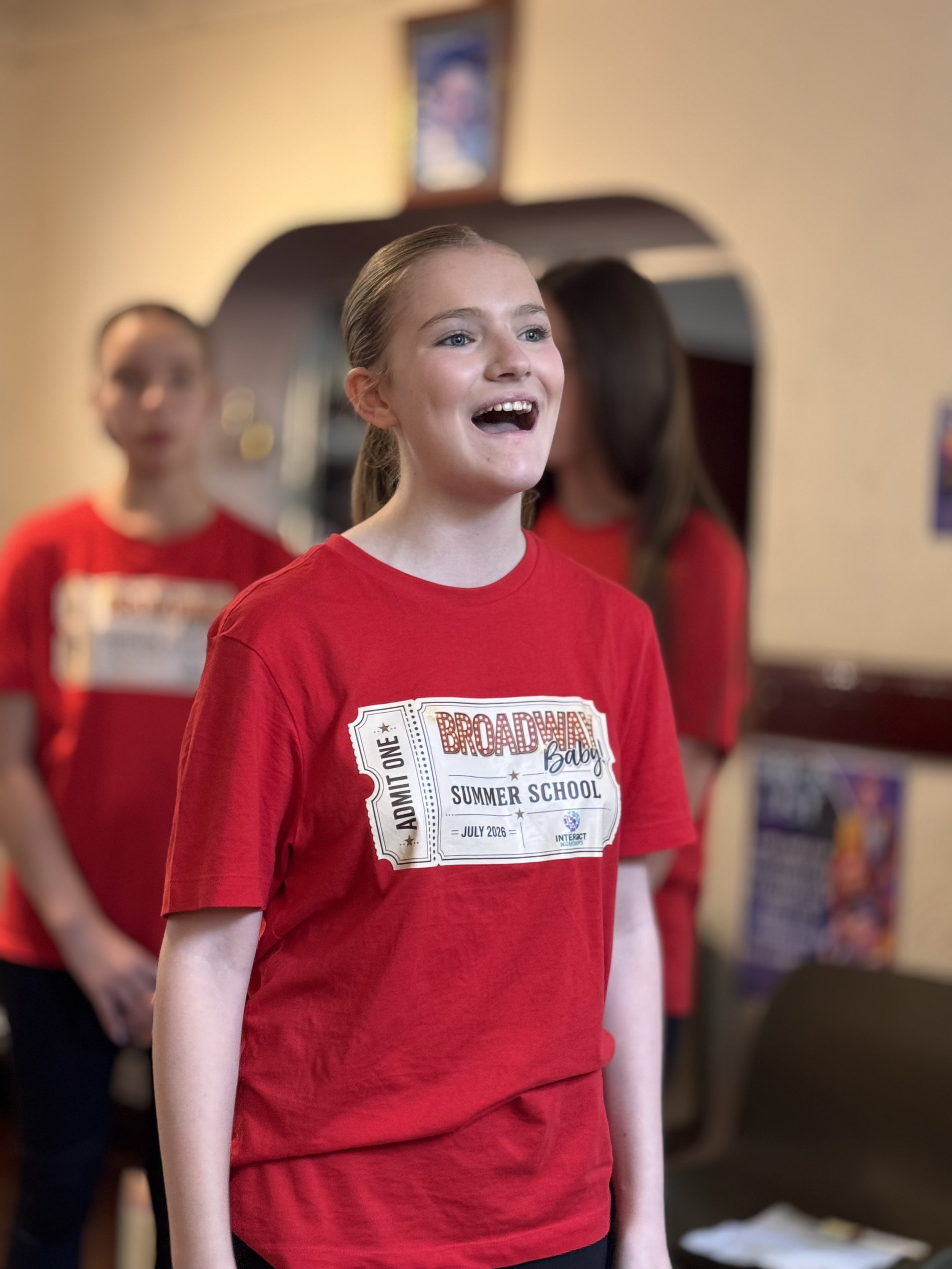 Close-up of a girl with blonde hair wearing a red T-shirt, smiling and looking ahead, with other children in similar red shirts in the background. The T-shirt has a graphic print that says 'Broadway Baby Summer School'.