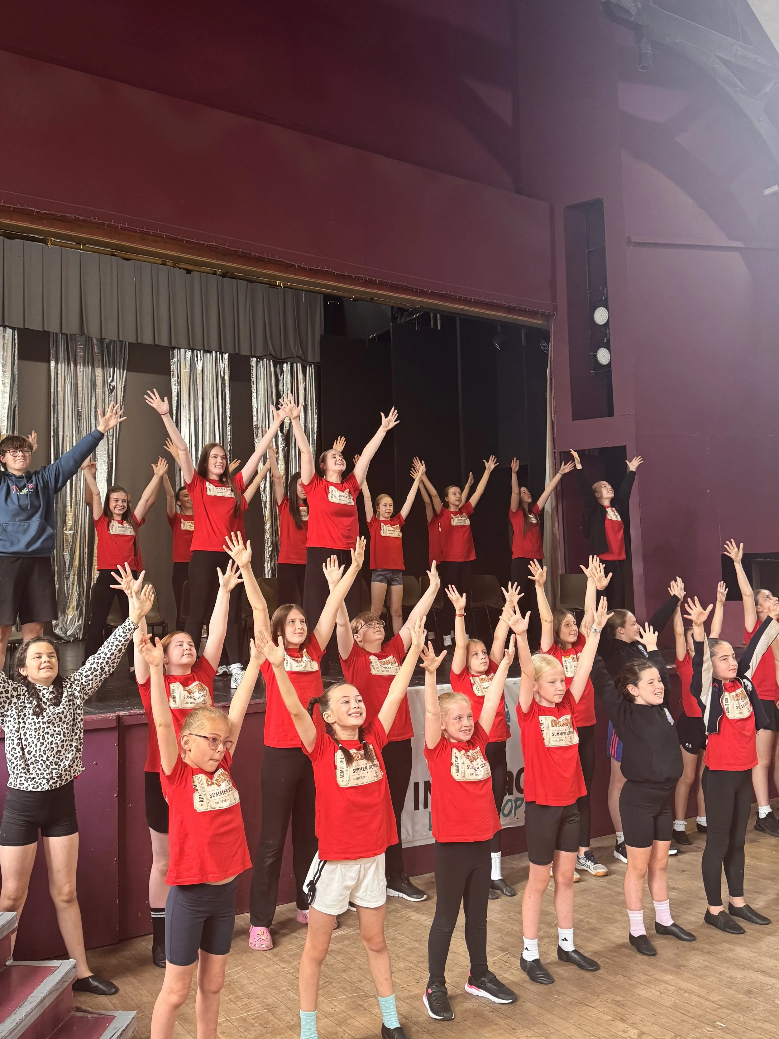 A group of children on a stage, raising their hands, in a dance or performance, with some adults around them, in a room with a curtain and stage lighting.