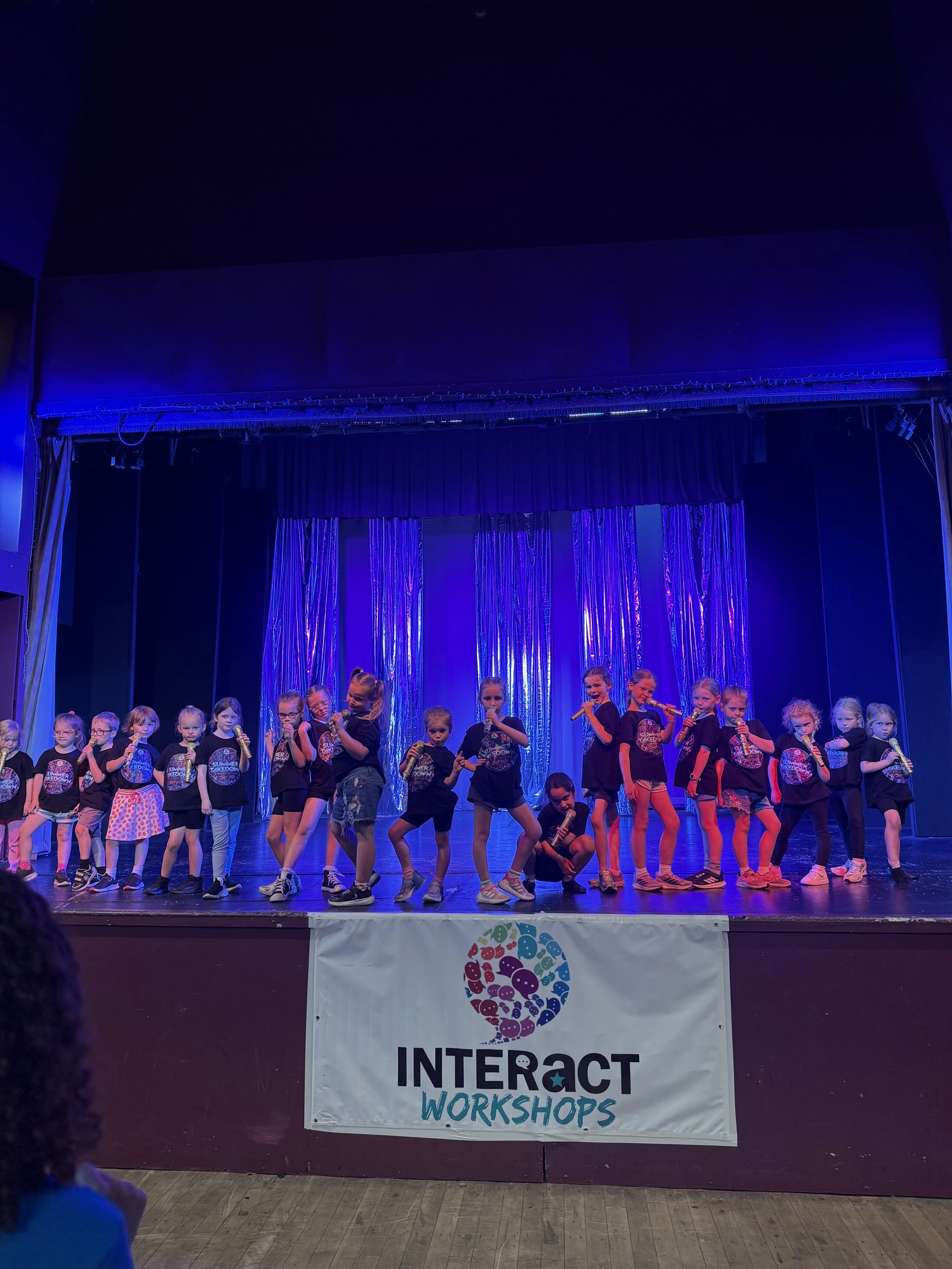 Group of children standing on stage during a workshop performance, with some holding microphones and smiling, in front of a blue curtain with metallic streamers, and a banner that reads 'Interact Workshop'.