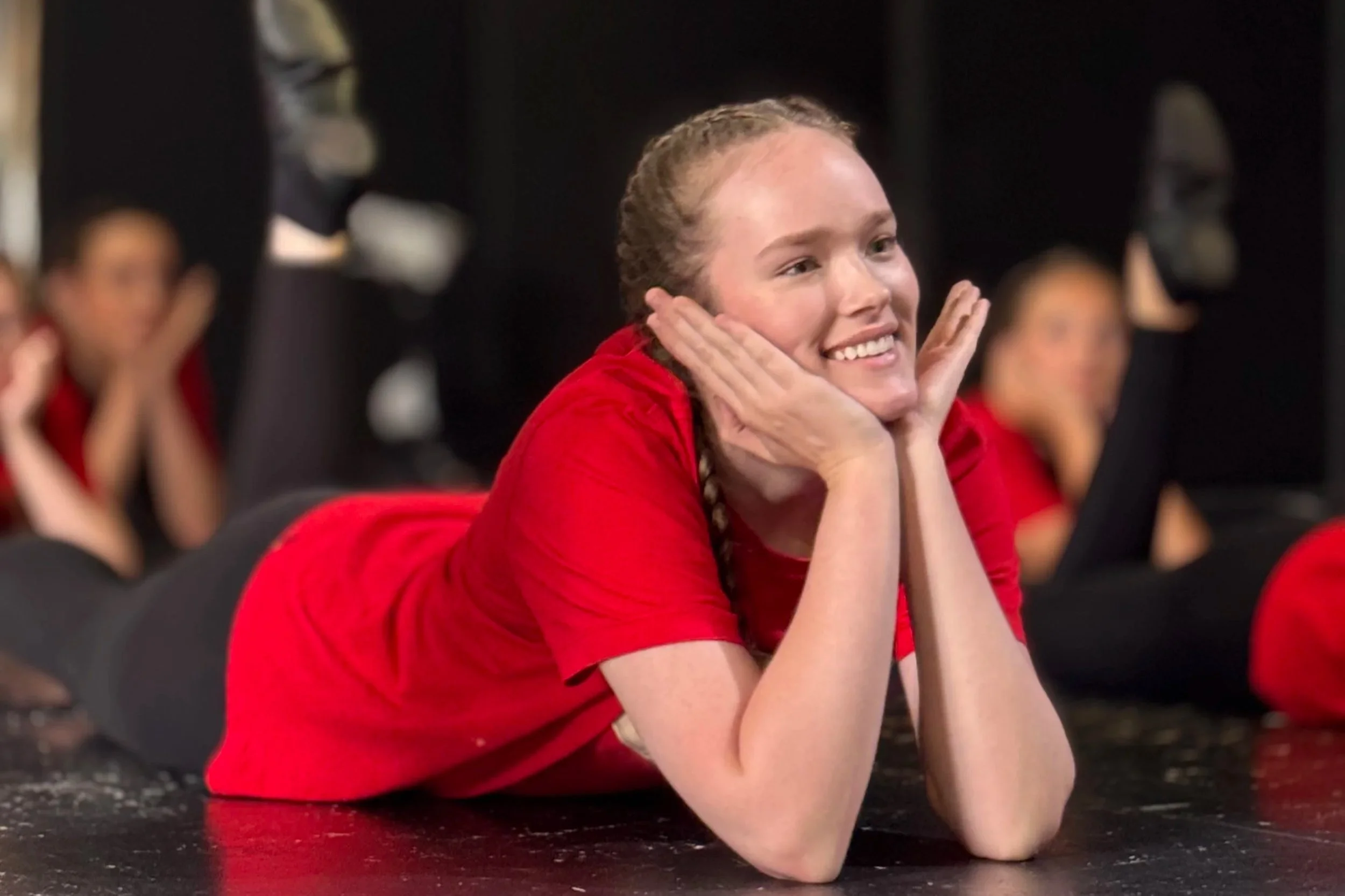 A young woman with braided hair and a red shirt resting her chin on her hands, smiling, sitting in a room with other women who are also in similar poses.