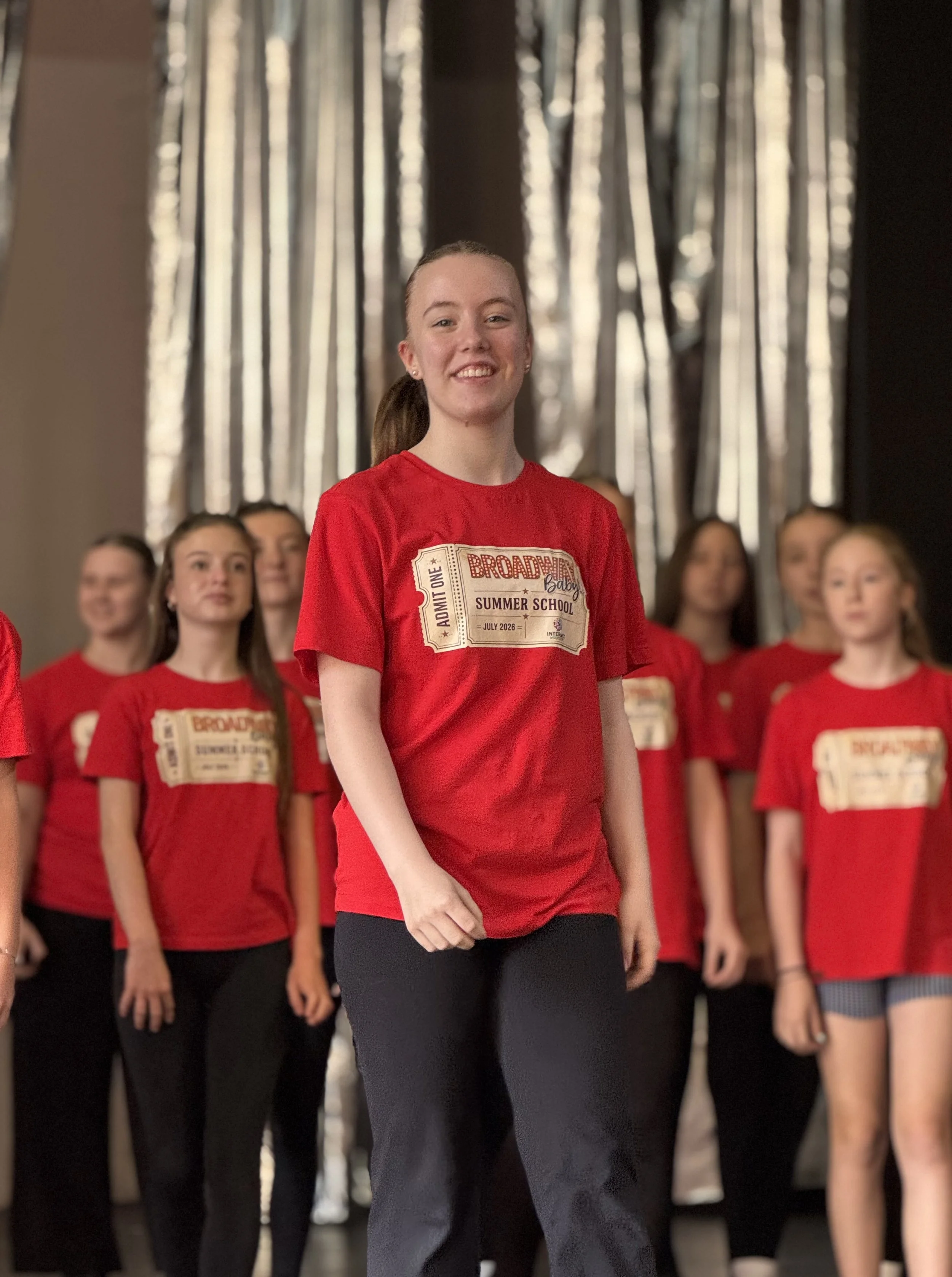 A woman with her hair tied back, wearing a red t-shirt with printing on it, standing in front of a group of women also dressed in red, black, or patterned clothing, against a background of vertical reflective curtains.