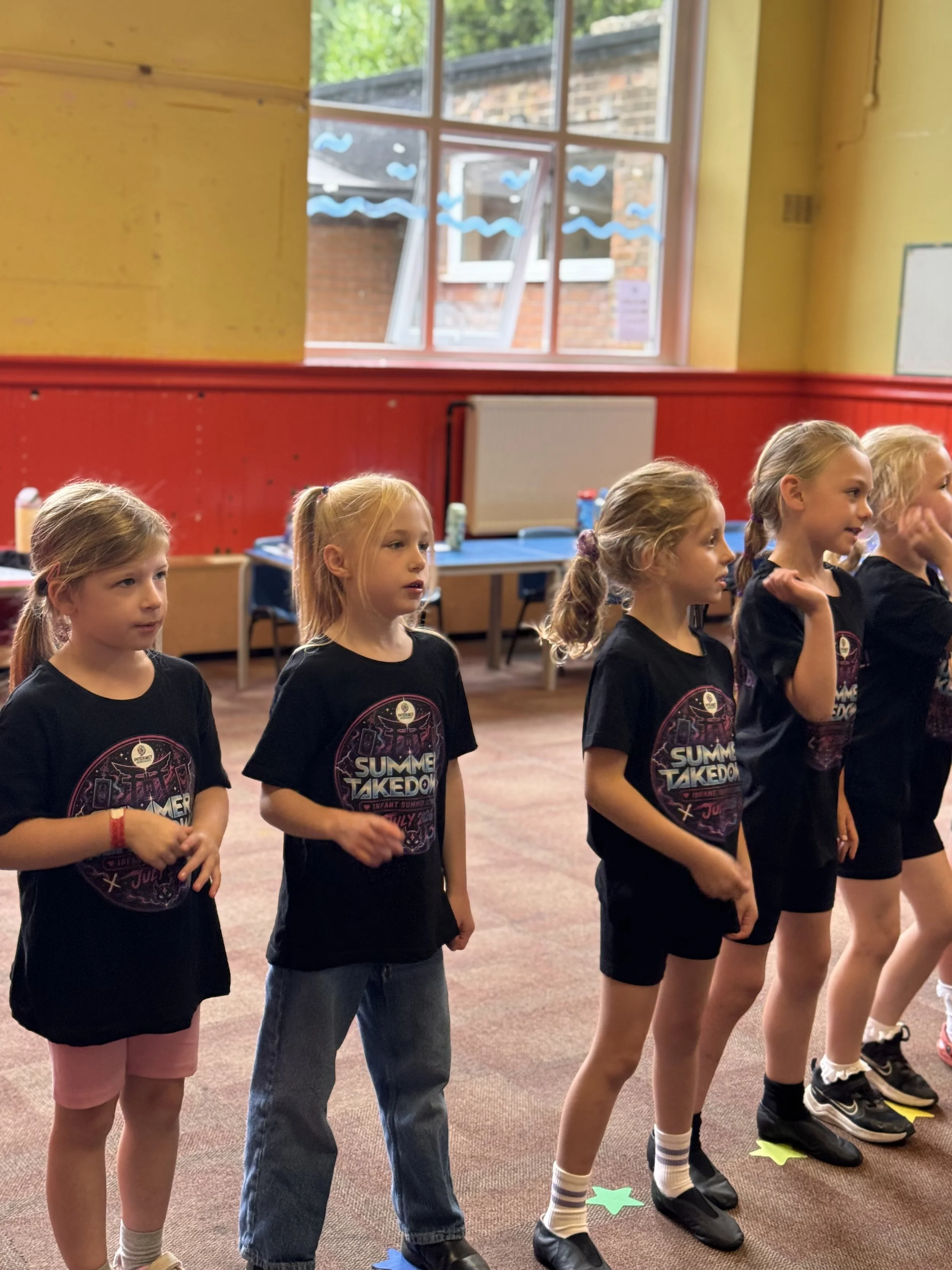 Group of young girls standing in a line indoors, wearing black t-shirts with a colorful logo, on a carpeted floor with colored star-shaped markers.