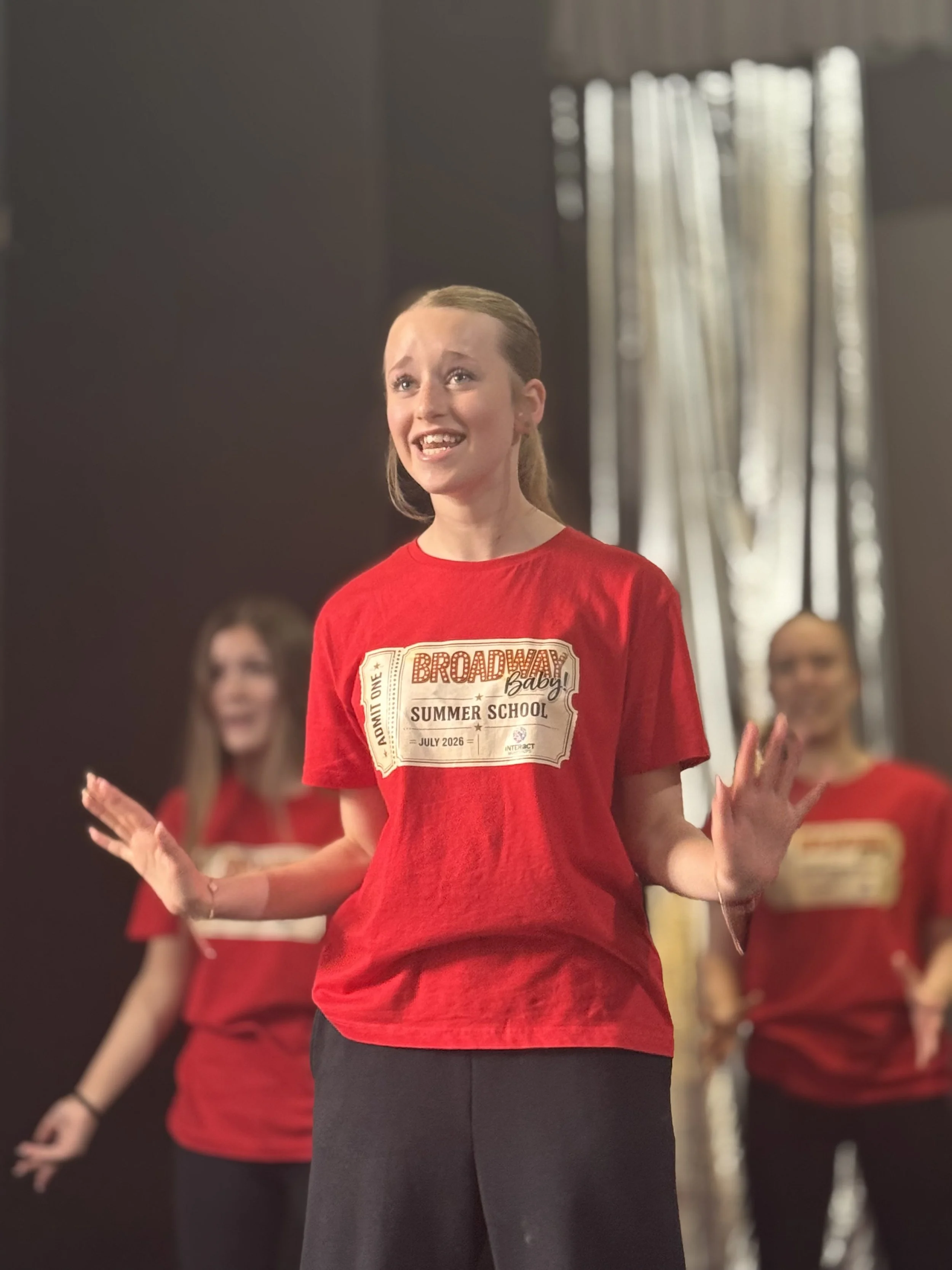 A young girl with blonde hair in a ponytail wearing a red t-shirt that says Broadway Baby Summer School, standing with her hands open, smiling, with other girls in red shirts blurry in the background.