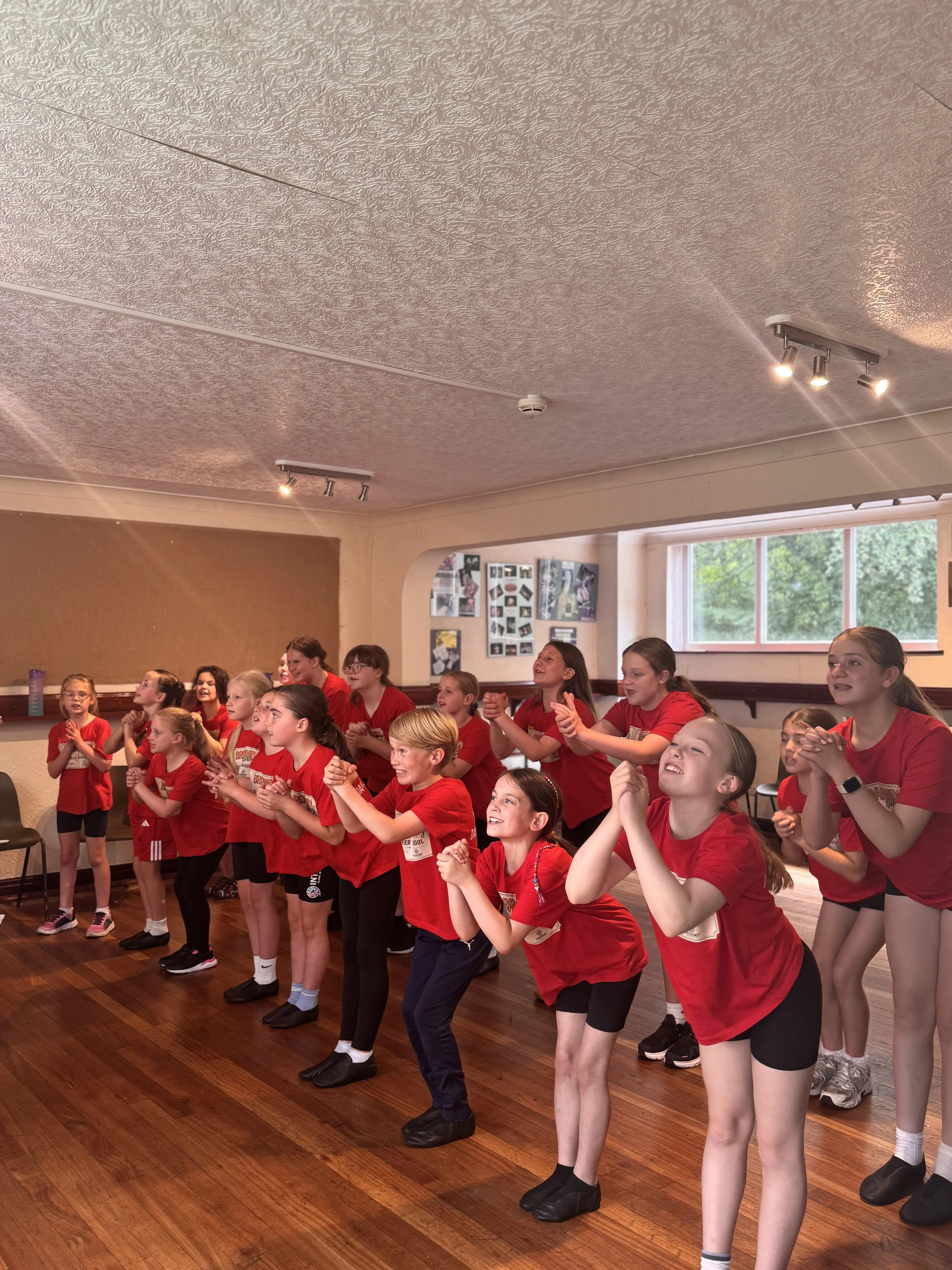 A group of children, mostly girls, wearing red T-shirts and shorts or leggings, are standing on a wooden floor in a room with a brown bulletin board and windows. They are actively engaged, smiling, and appear excited or happy.