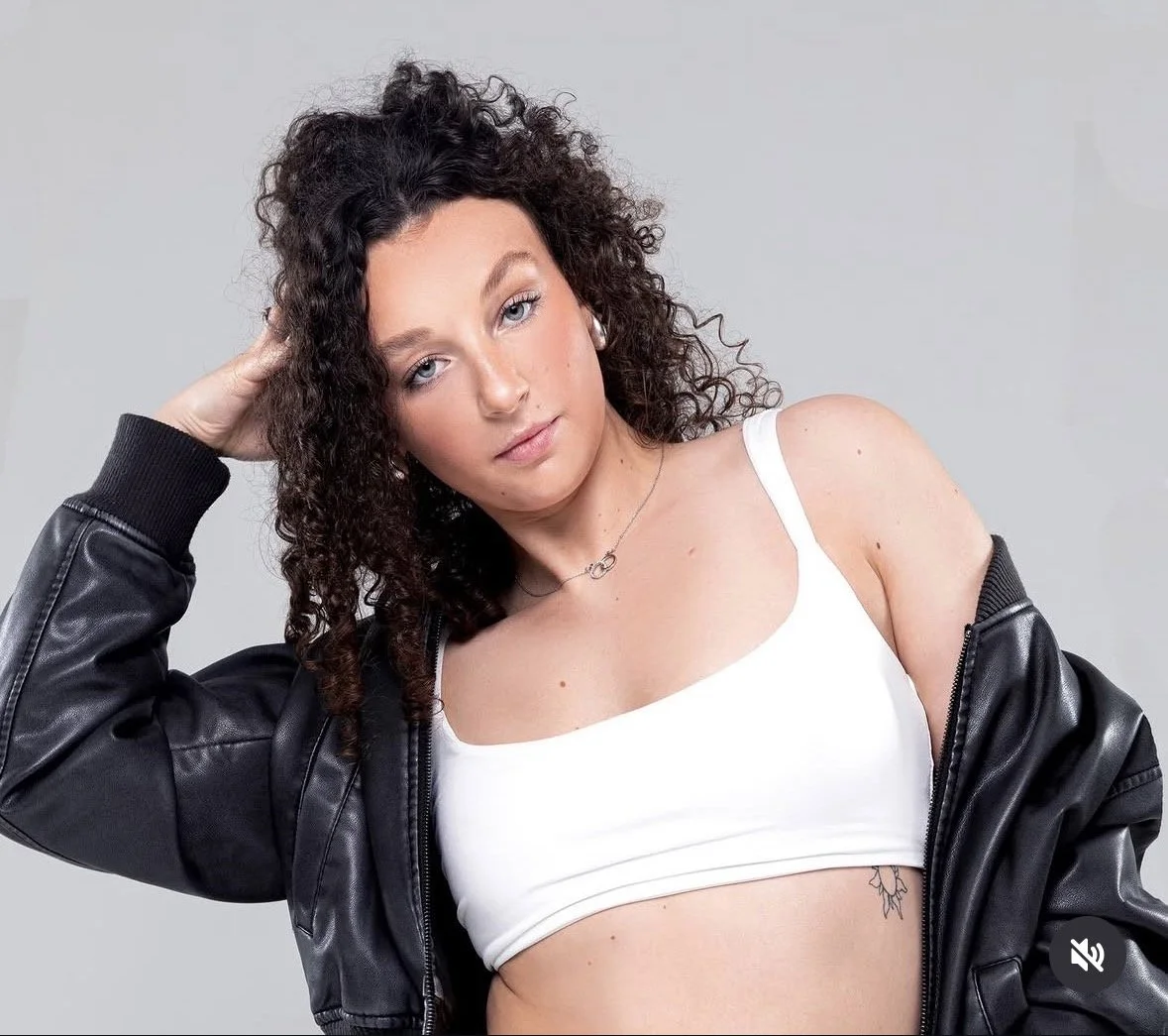 A young woman with curly dark hair, wearing a white crop top and a black leather jacket, posing with her hand on her head against a plain gray background.