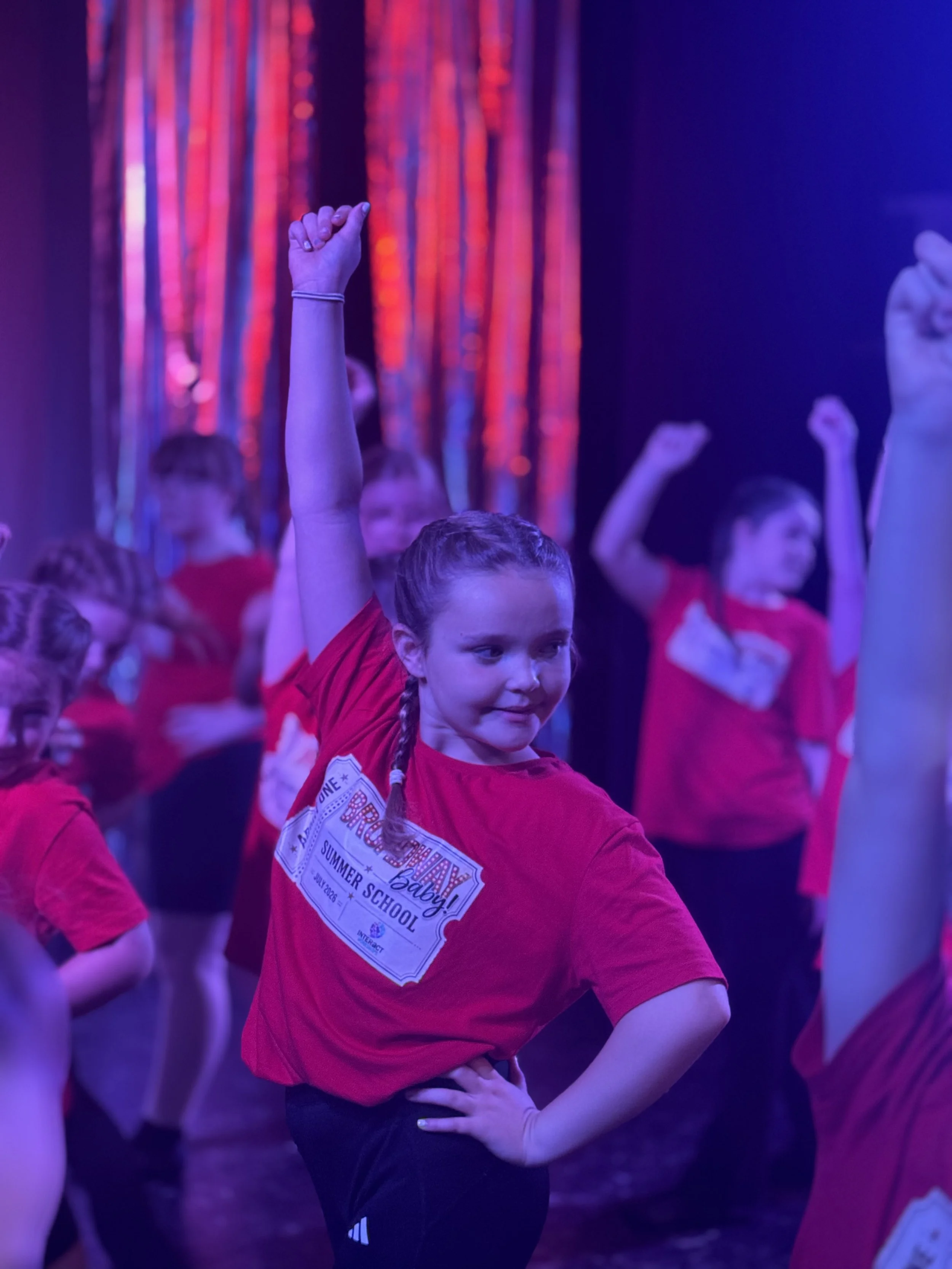 Young girl participating in a dance or exercise class with other children, raising her arm, in a room with vibrant colorful lighting and curtains.