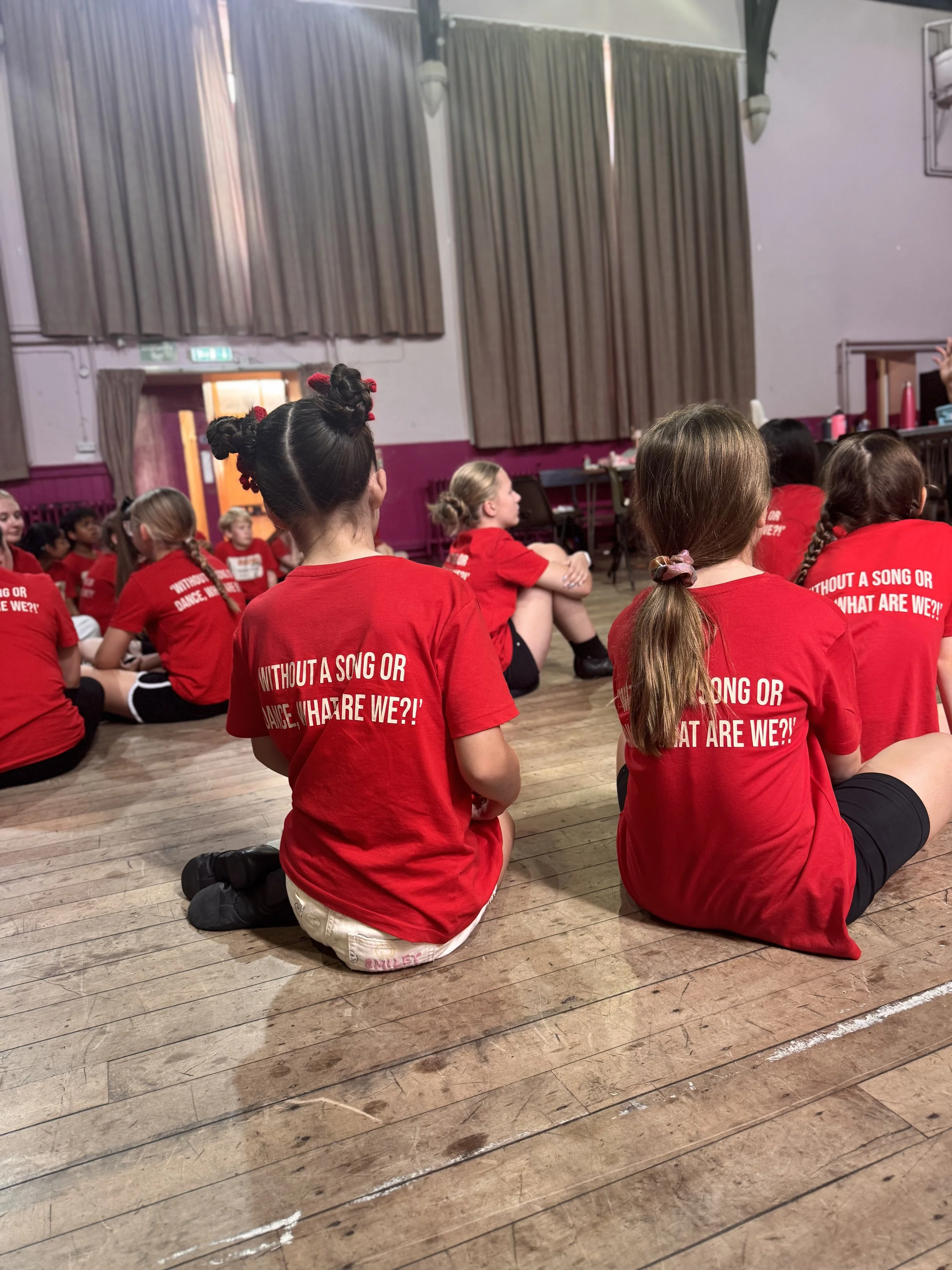 Children sitting on a wooden floor in a large room, facing forward, wearing red T-shirts, participating in a group activity or performance.