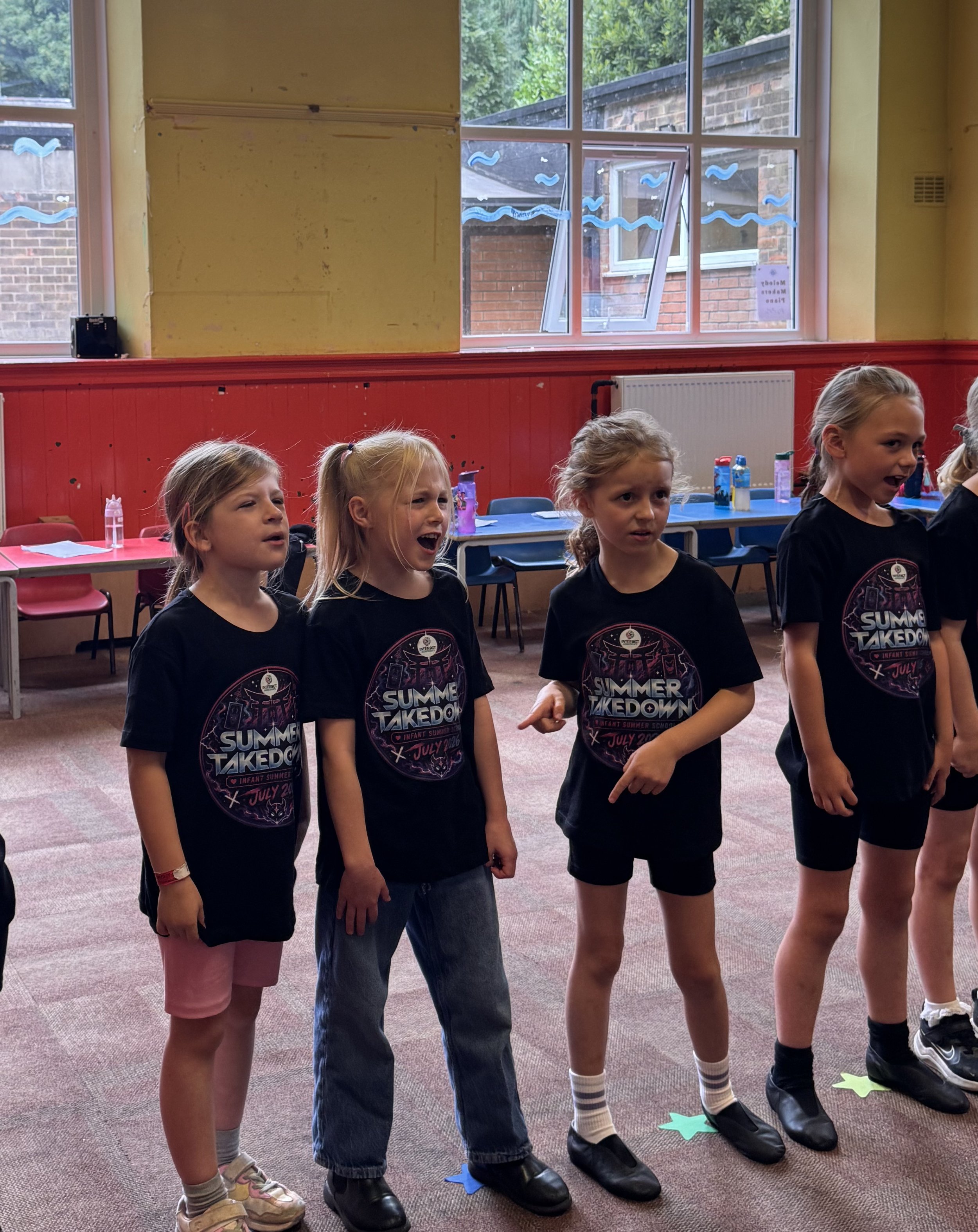 Four young girls standing in a row indoors, wearing black T-shirts with a 'Summer Takedown' logo, and standing on colored paper shapes.