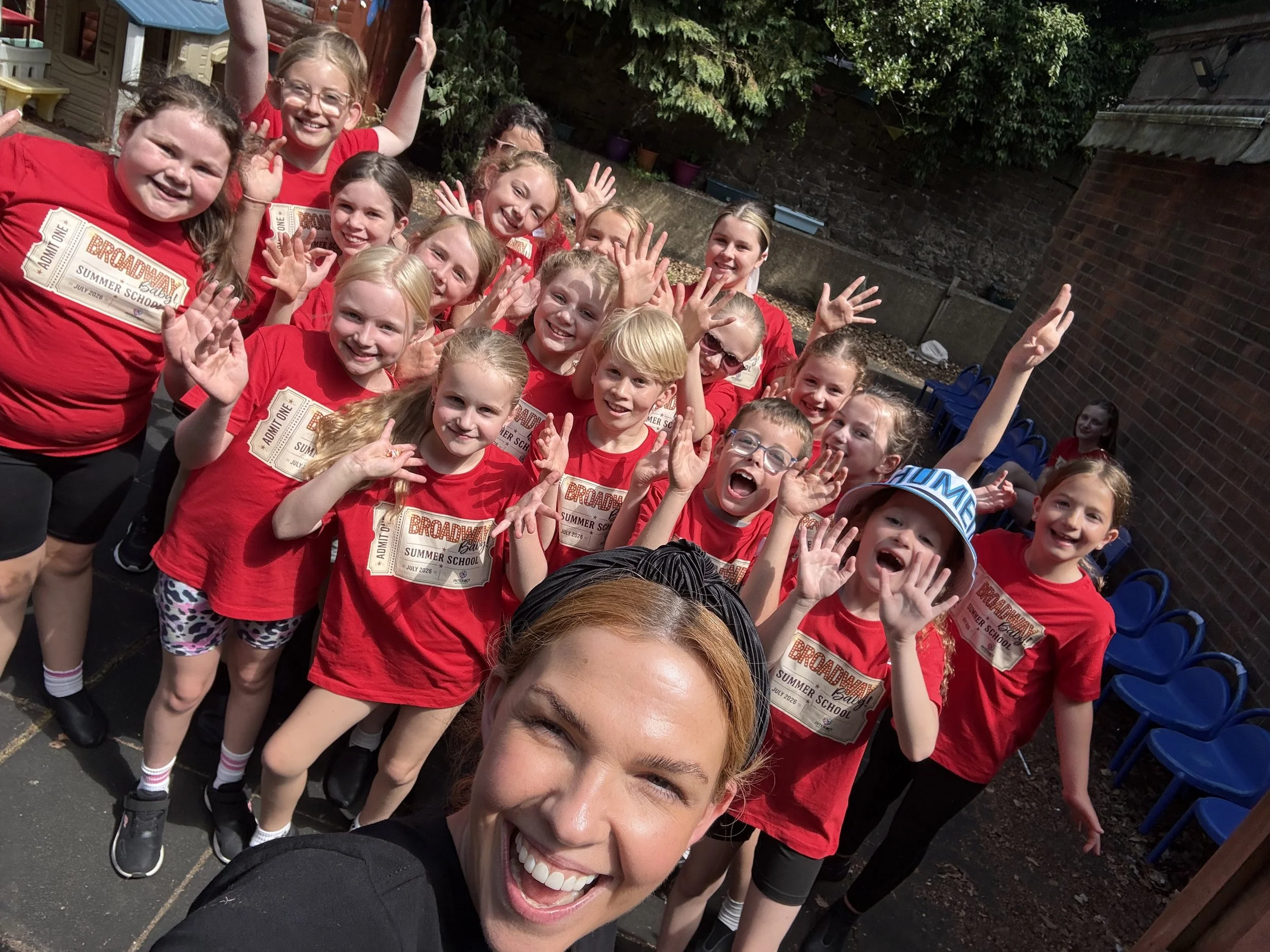 A group of children and one adult woman smiling and waving at the camera. The children are wearing red t-shirts with a summer school logo, and the woman is smiling broadly. They are outdoors, possibly in a backyard or playground, with trees, bushes, 