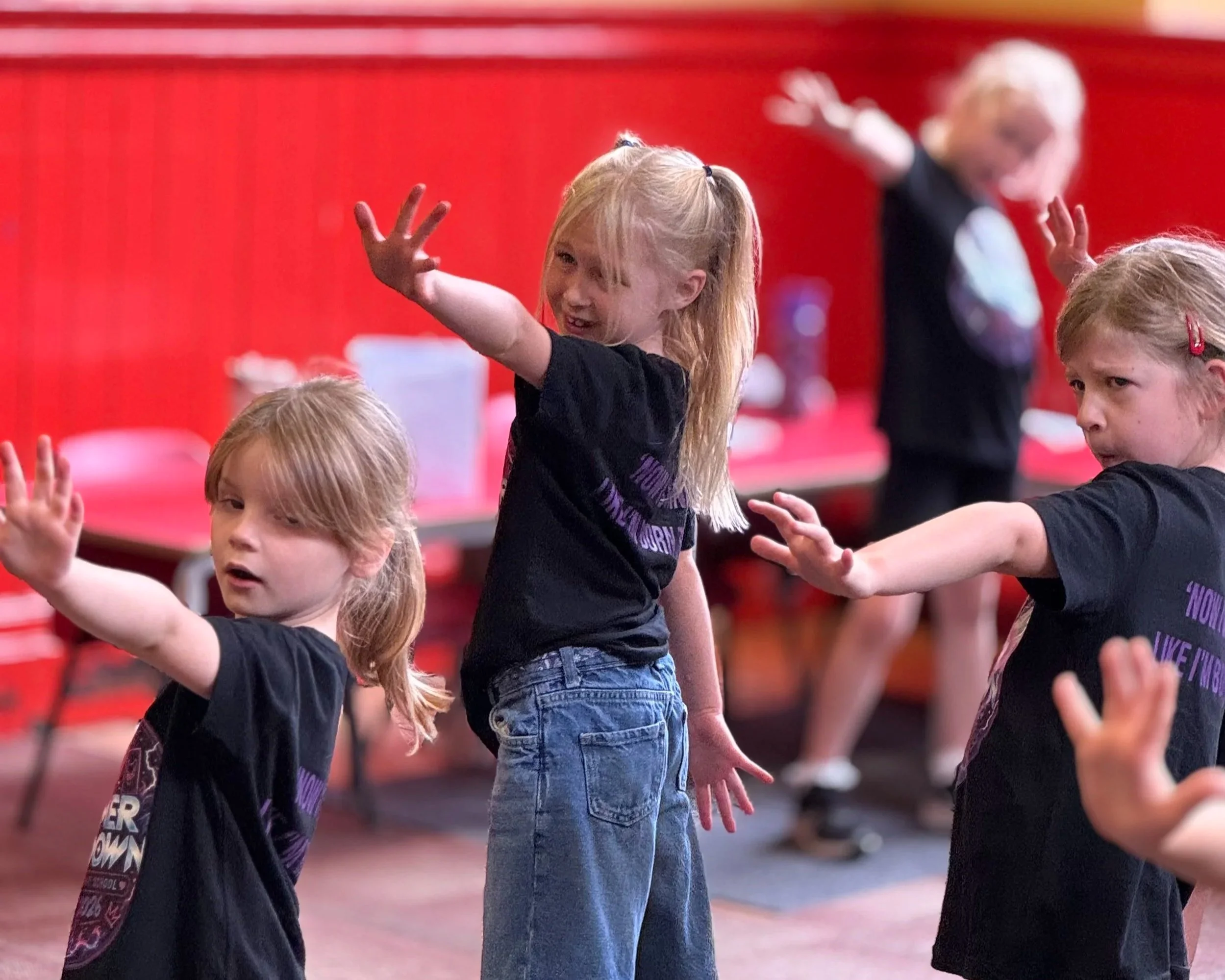 Children participating in a dance or exercise class, with one girl at the center raising her hand and others around her, against a red background.