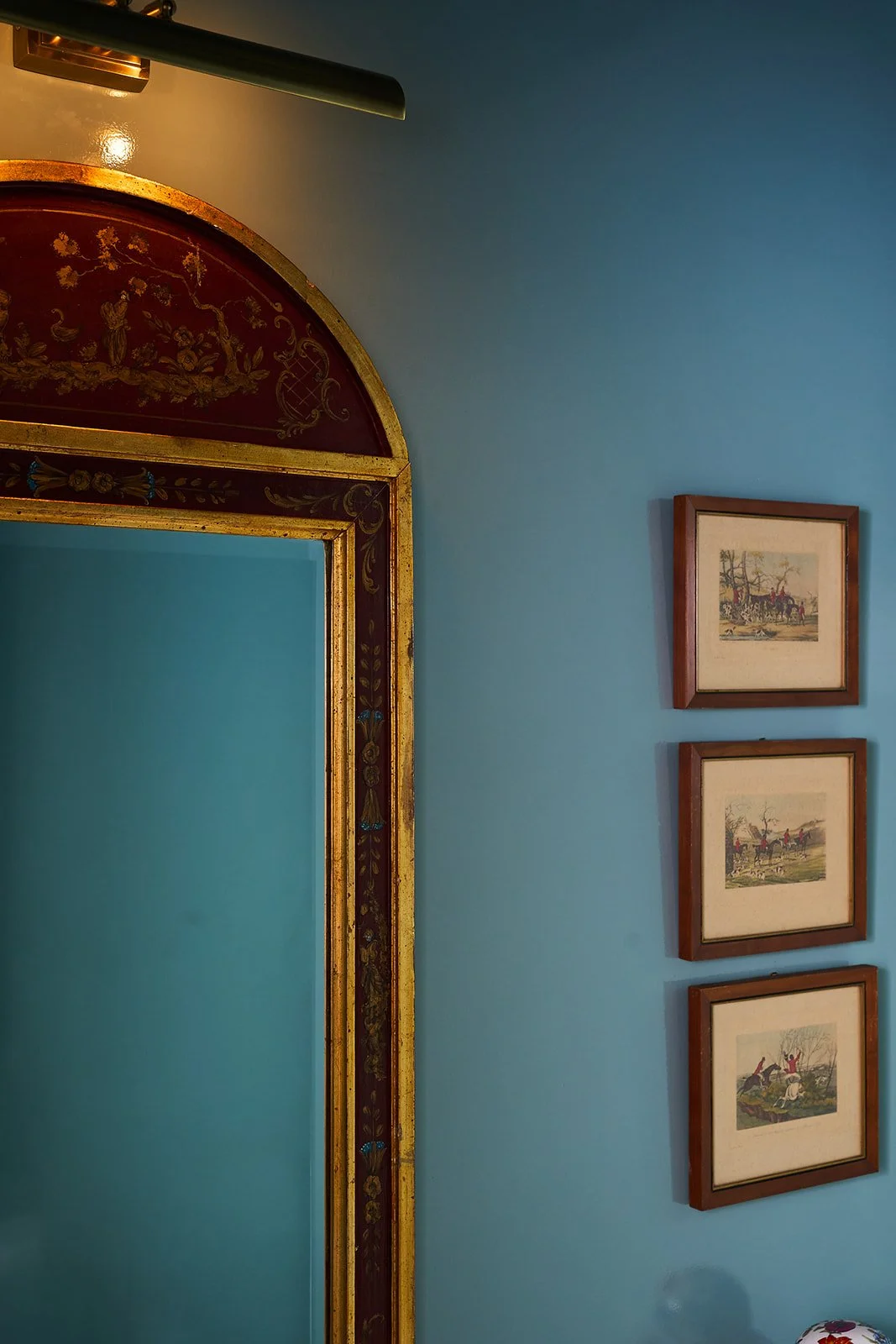 Part of a room with a blue wall, three framed pictures of horse-drawn carriages, and a decorative mirror with a gold and maroon ornate frame.