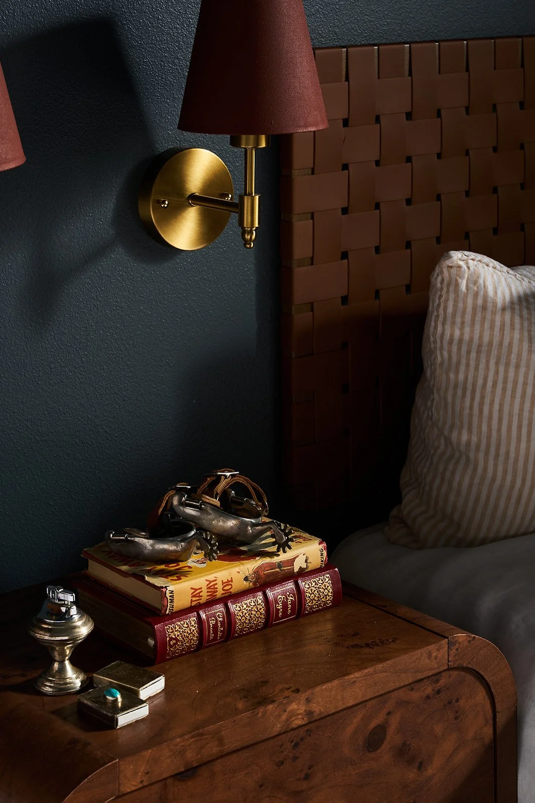 Close-up of a bedroom nightstand with books, decorative shoes, a small silver vase, and a box. A wall-mounted brass sconce with a red lampshade is above the nightstand. Part of a wooden headboard and a striped pillow are visible.
