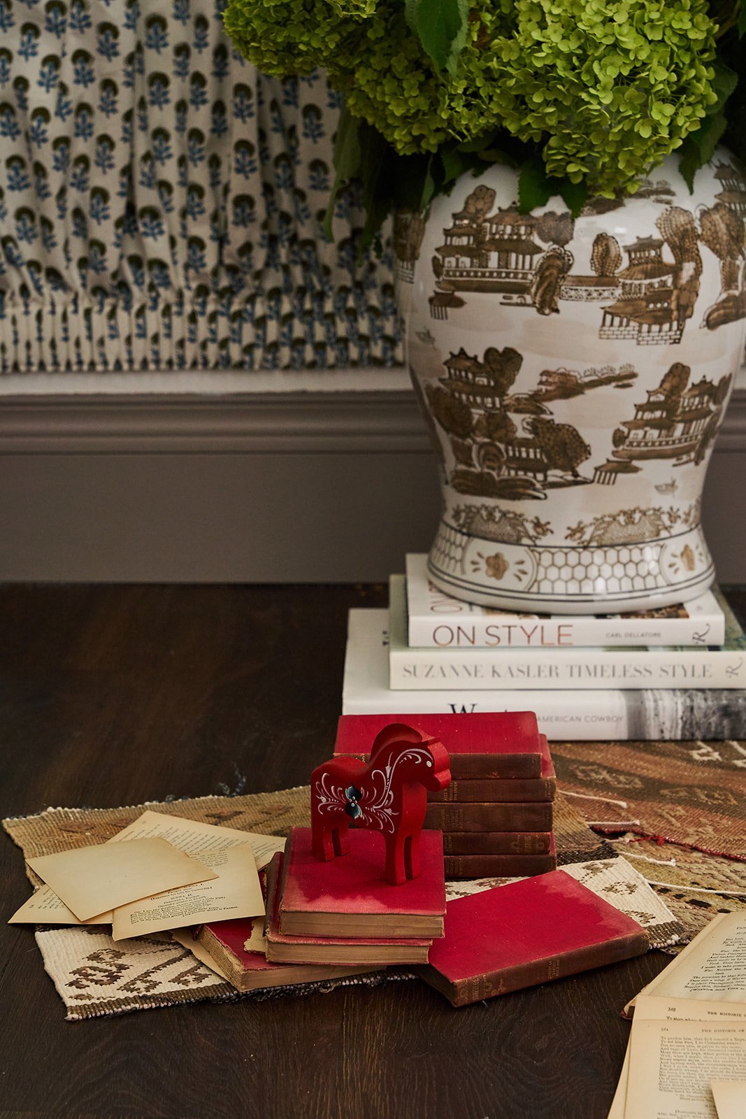 Decorative arrangement on a table featuring a large ceramic vase with Asian-themed illustrations, green hydrangea flowers, books, a red wooden horse figurine, vintage books, and papers. In the background, a patterned fabric with blue and white design
