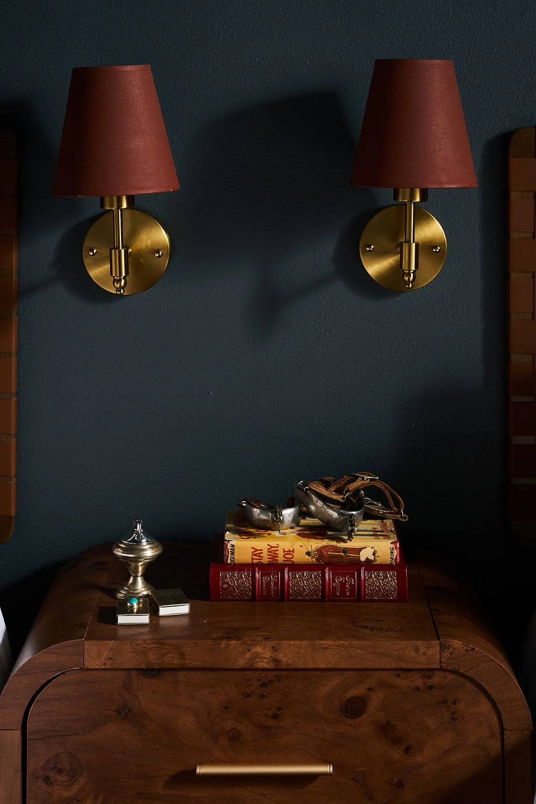 Two wall-mounted lamps with brown shades and brass fixtures above a wooden side table. The table holds a stack of books, a decorative silver box, and a pair of sunglasses.