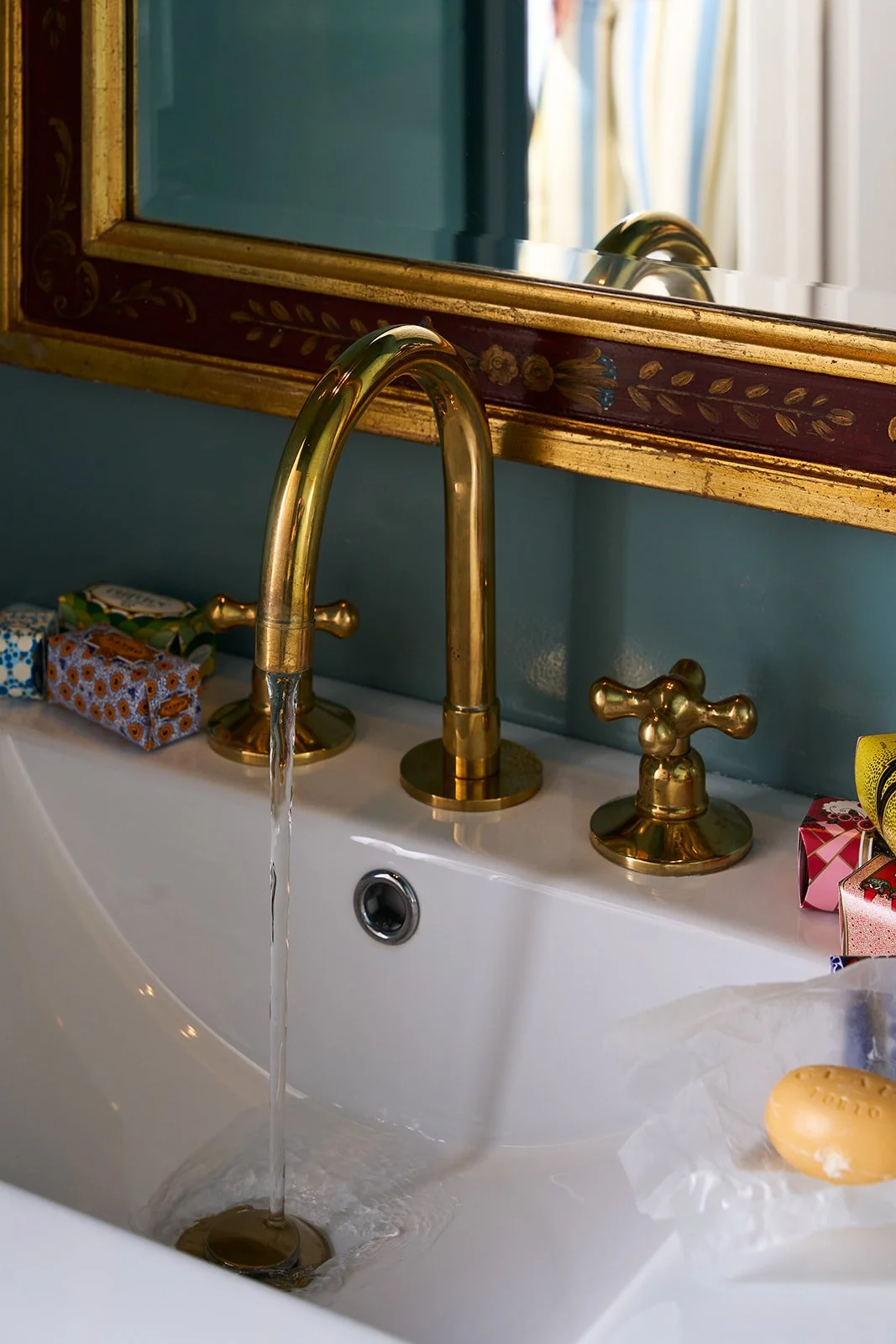 A white bathroom sink with a gold faucet and handles, with water running from the faucet. There are colorful soap bars and gift boxes on the sink. A large mirror with a decorative gold and maroon frame is above the sink, reflecting a window with curt