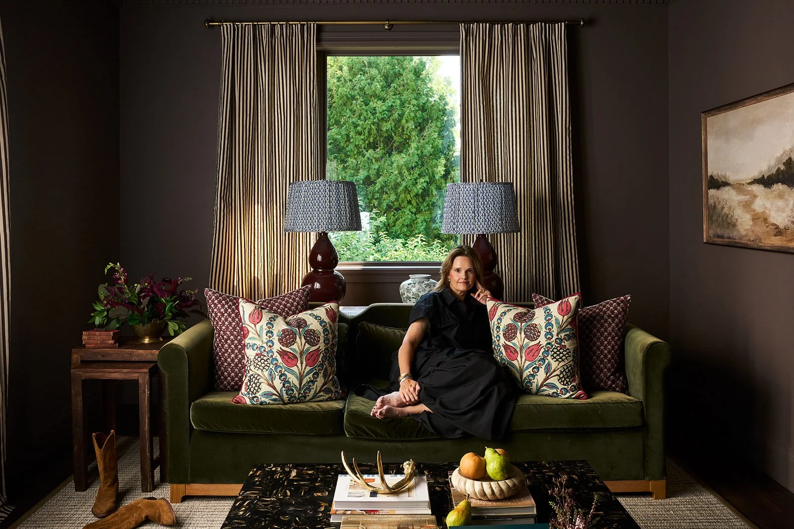 Woman sitting on a green velvet sofa in a cozy living room, surrounded by patterned pillows, with a window showing greenery outside, flanked by striped curtains, and a wooden side table with a flower arrangement on the left.