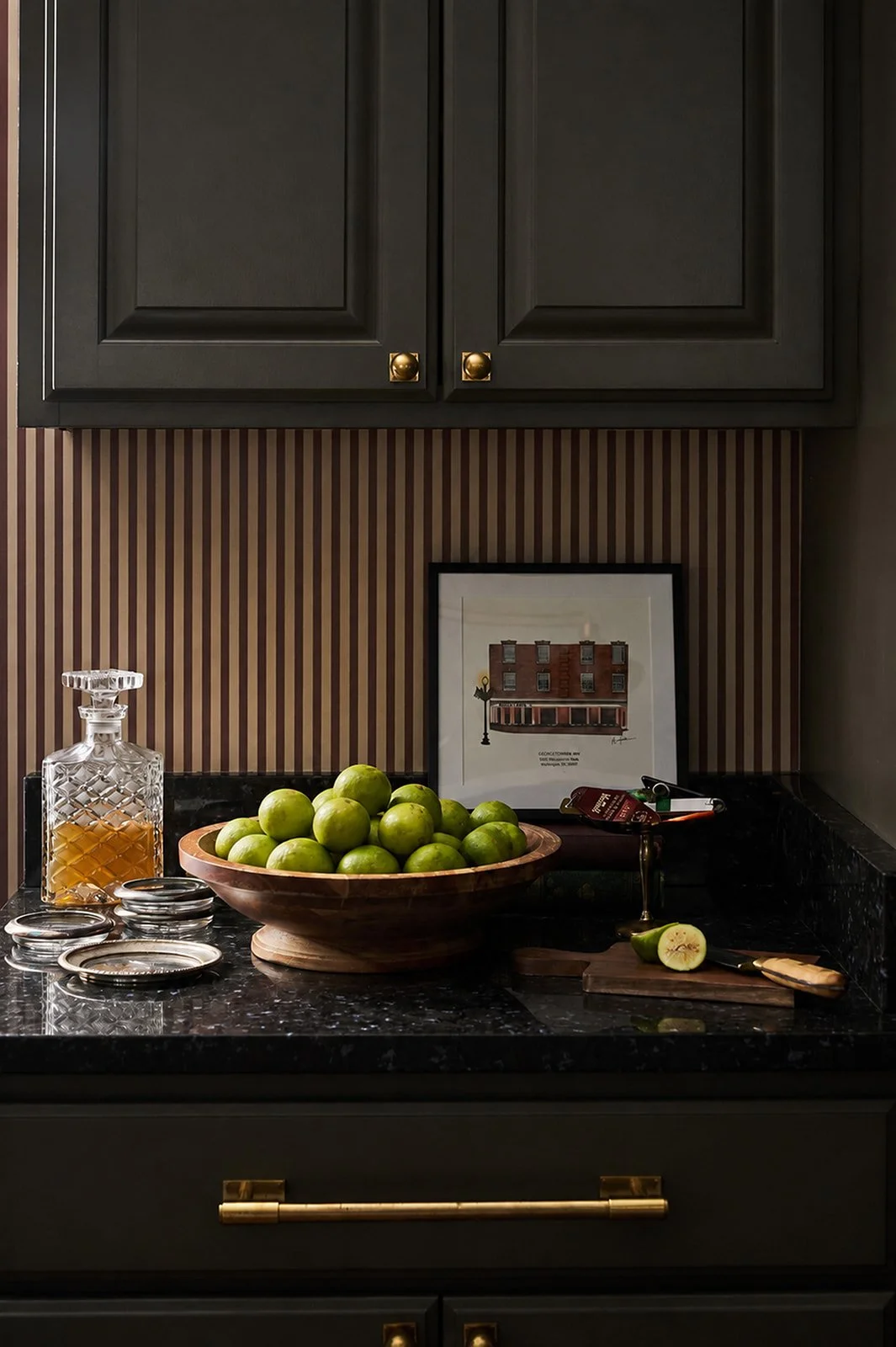 A kitchen countertop with a wooden bowl of green limes, a glass decanter, a framed picture of a building, a small tray with jars, and a sliced lime with a knife on a cutting board.