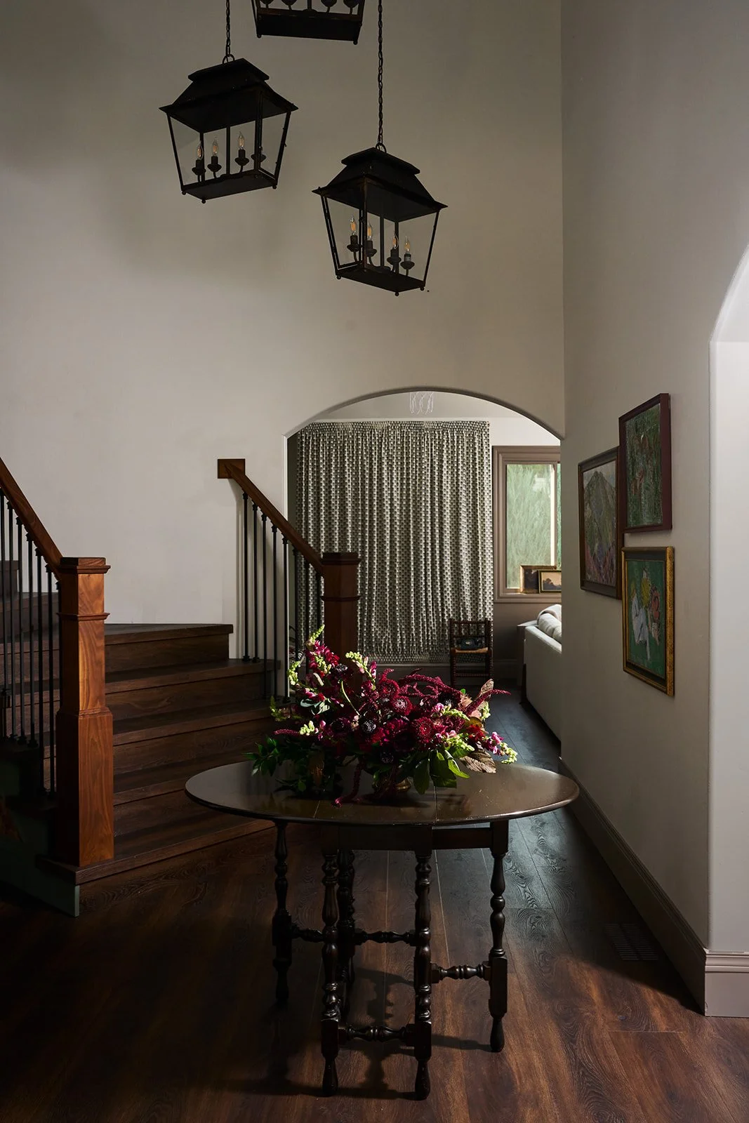 Entryway with wooden stairs, a round wooden table with a colorful flower arrangement, framed artwork on the wall, and a window with patterned curtains, illuminated by vintage hanging lanterns.